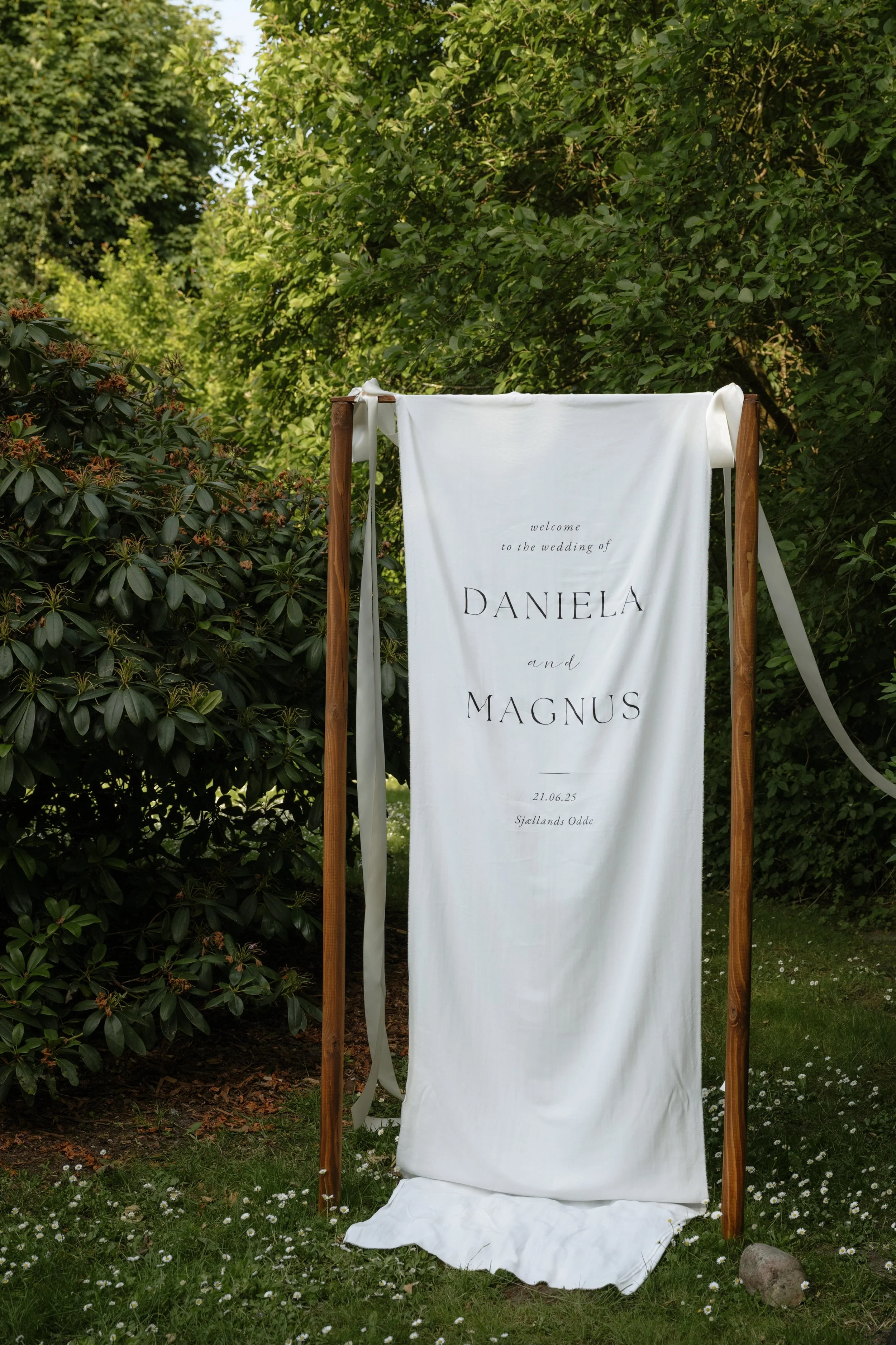 A white wedding sign with black text, hanging from a wooden frame outdoors surrounded by green trees and bushes, with small white flowers on the grass at the bottom.