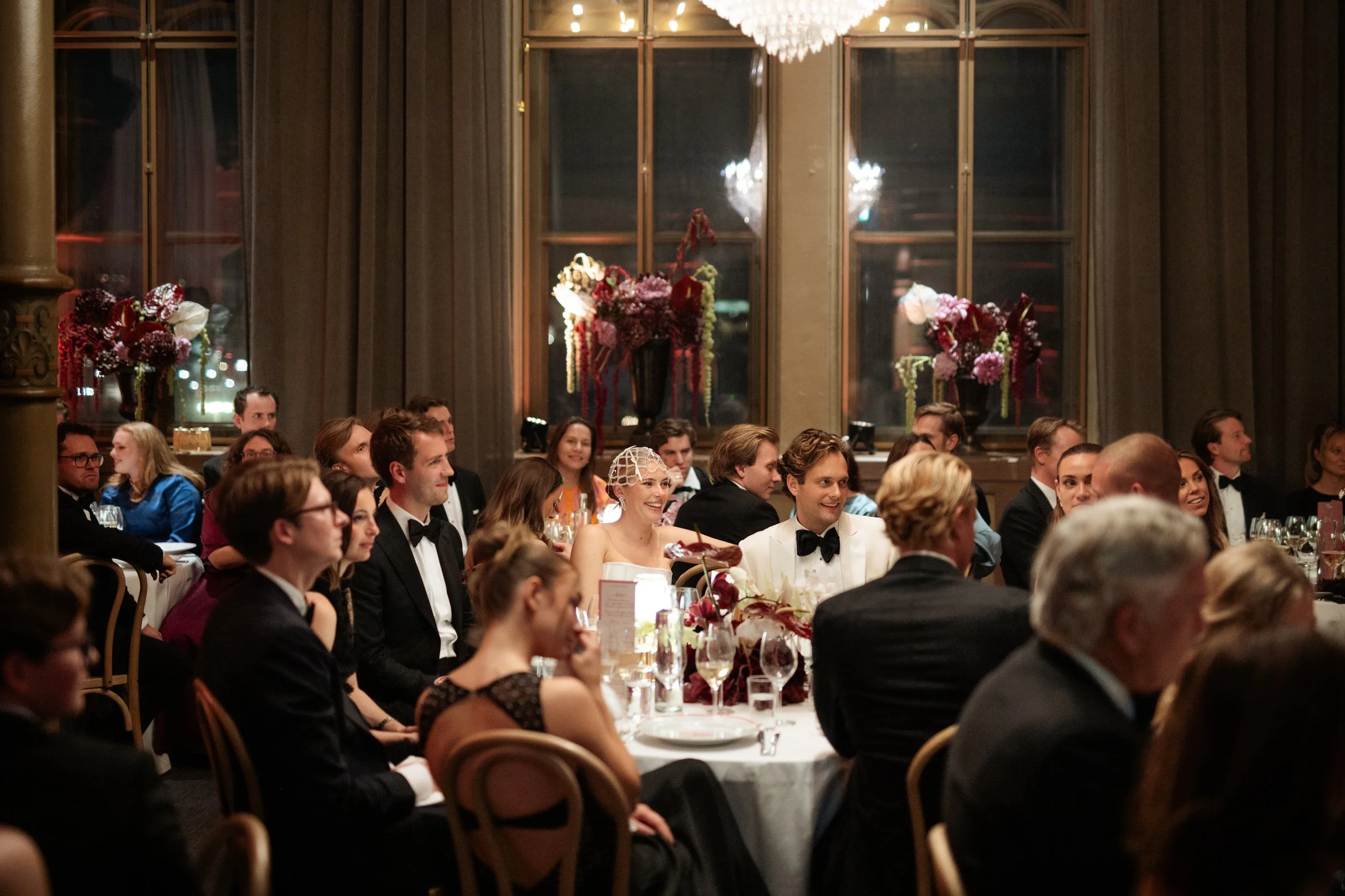Guests dressed in formal attire, including tuxedos and evening gowns, seated at tables with white tablecloths and floral centerpieces, attending a celebration or wedding reception in an elegant venue with large windows and floral decorations.