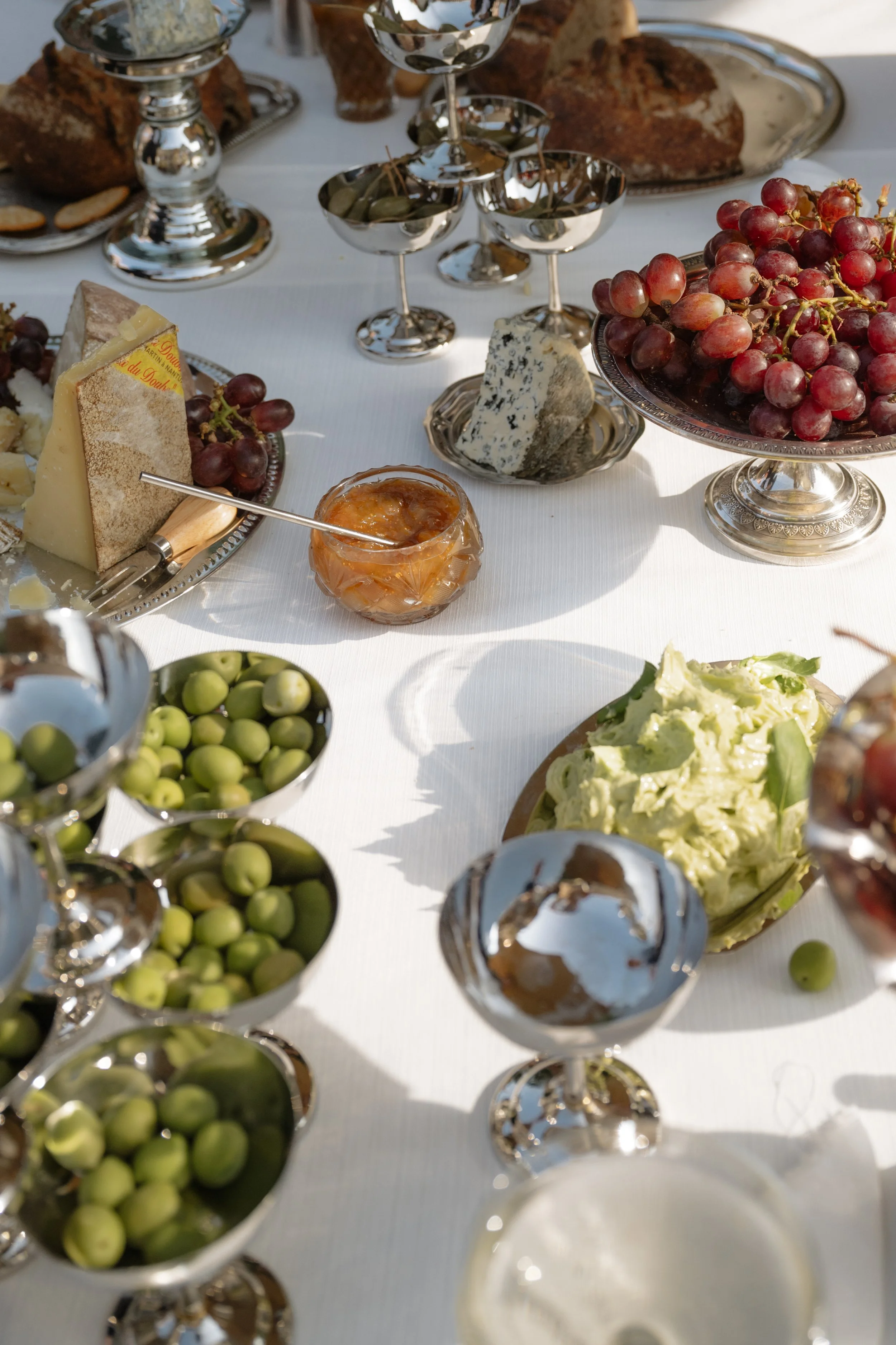 A table set with various cheeses, grapes, green olives, and a dish of guacamole, with silver goblets and a jar of jam or marmalade, likely for a meal or celebration.