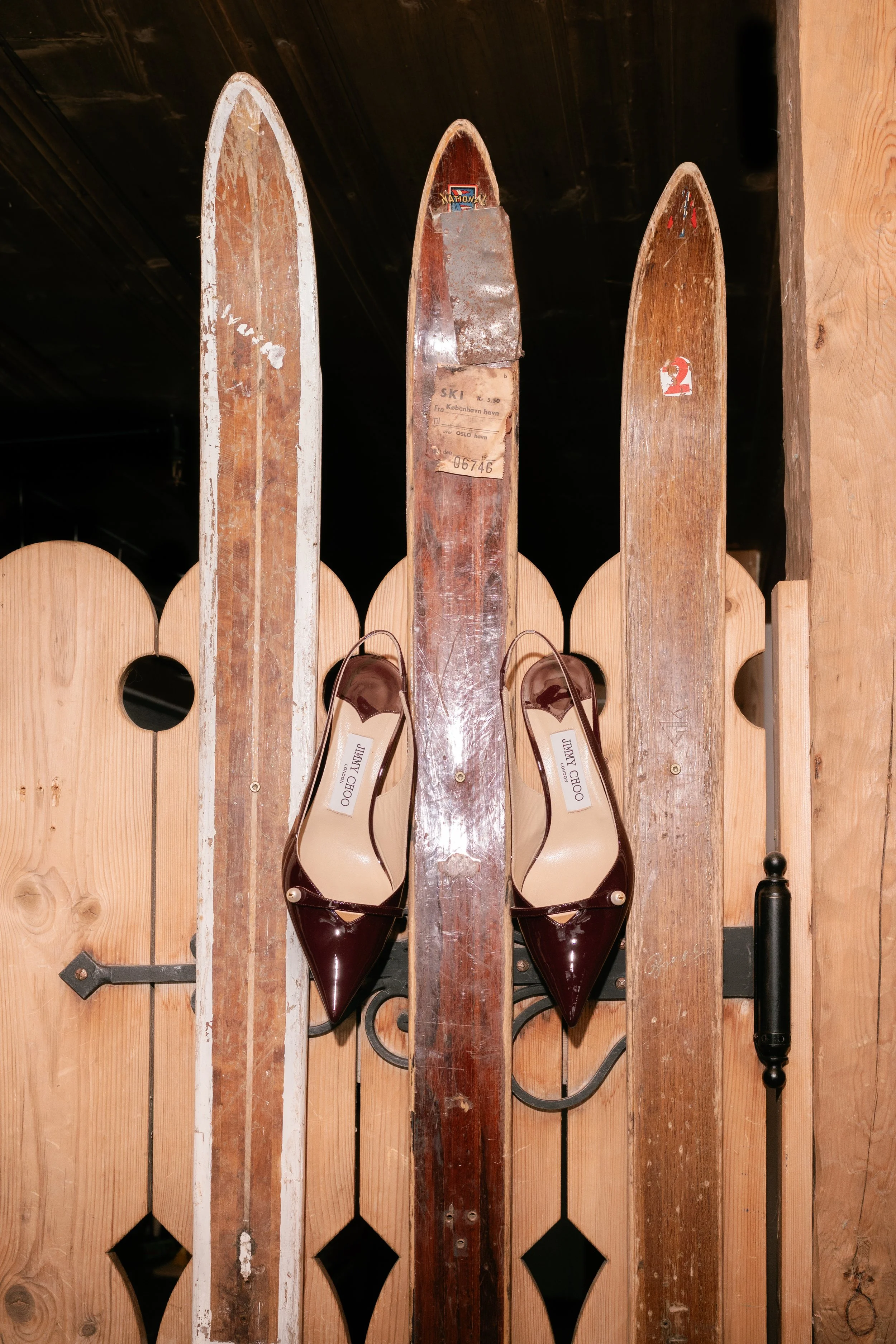 A pair of burgundy Jimmy Choo high heels hung on an old vintage pair of skis mounted on a wooden wall.