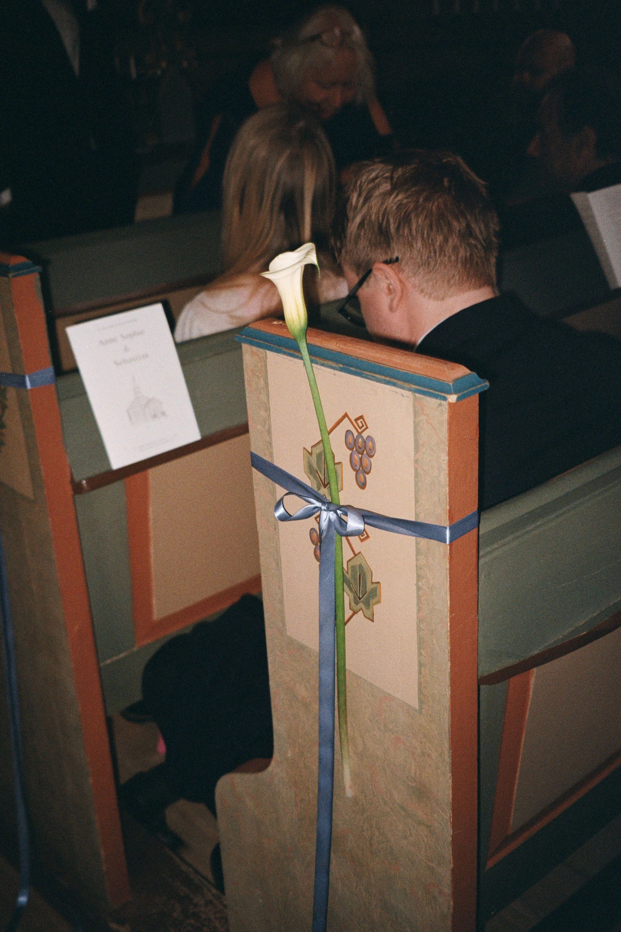 A white calla lily flower tied to a church pew with a blue ribbon, with wedding guests seated in the background.