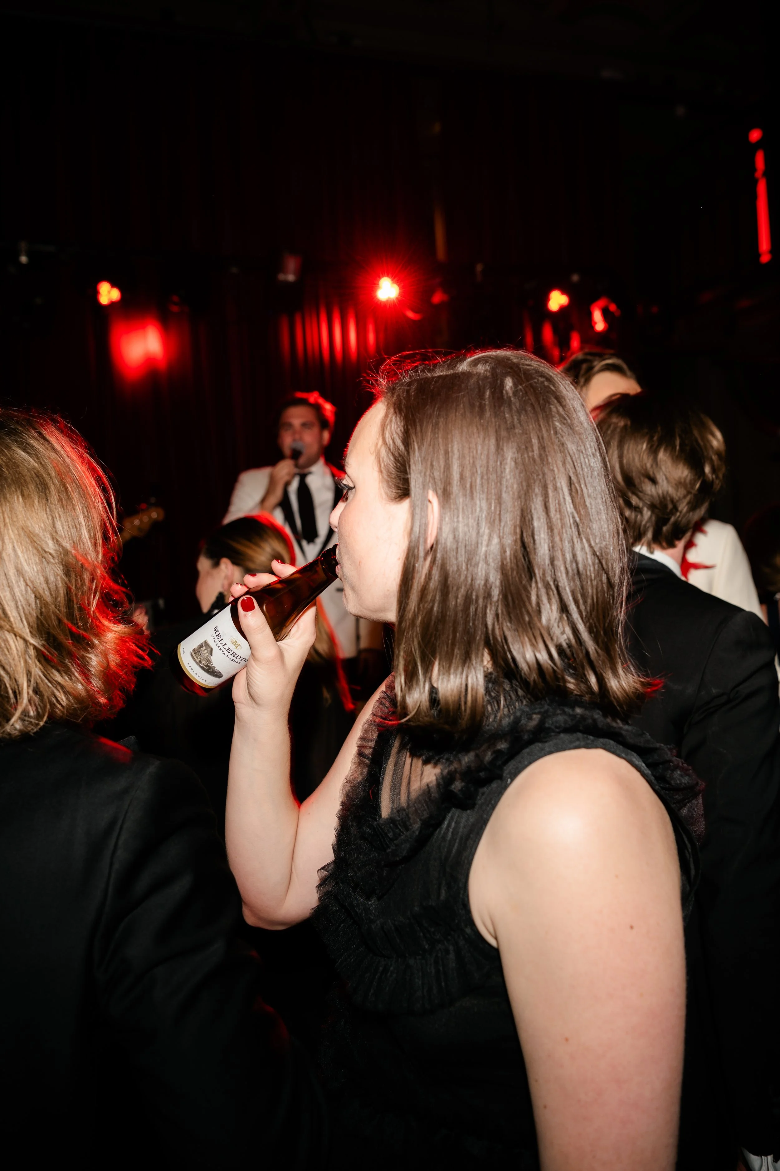 A woman with short brown hair wearing a black sleeveless dress is drinking from a beer bottle at a crowded party or nightclub with red lighting. Other people are visible around her, and a live performer with a white suit and black tie is in the backg