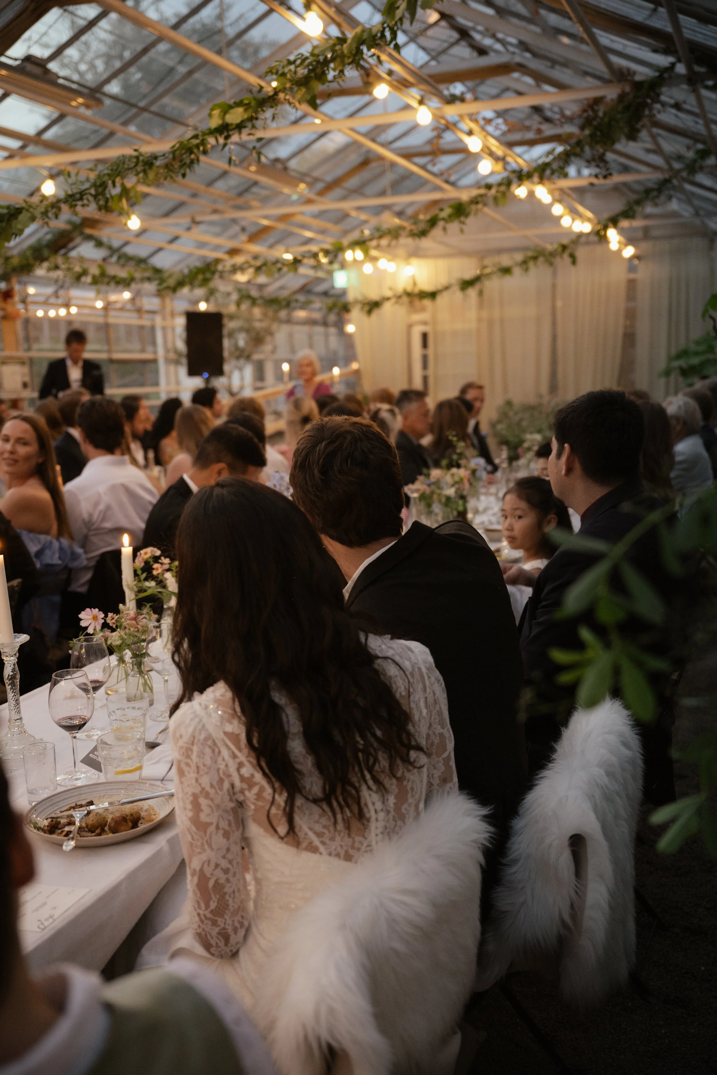 Guests seated at a long dinner table in a glass-roofed venue decorated with string lights and greenery, celebrating at a formal event or wedding reception.