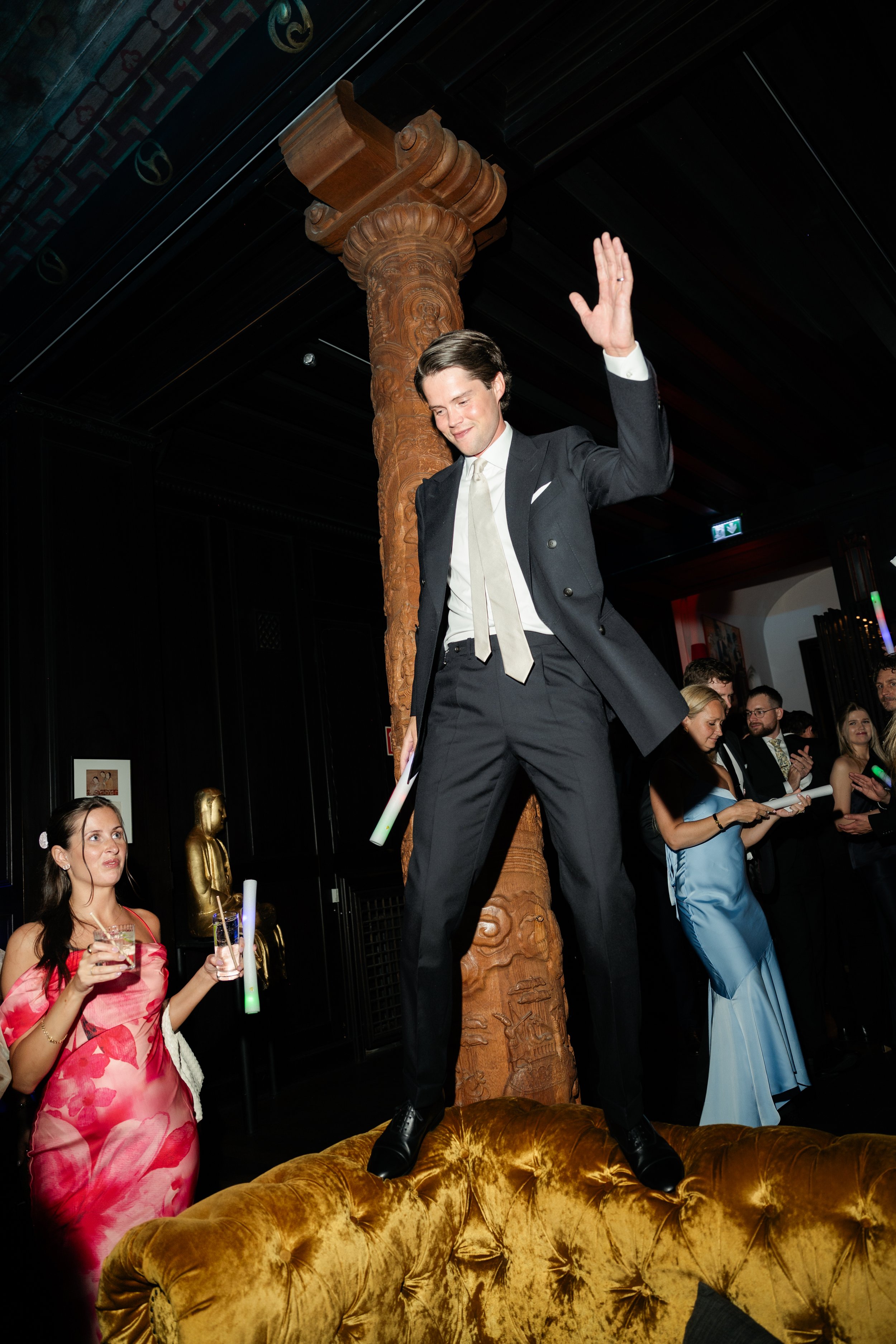 Man in a suit standing on a gold-colored couch, smiling and raising his hand, at a party with people holding drinks and socializing.