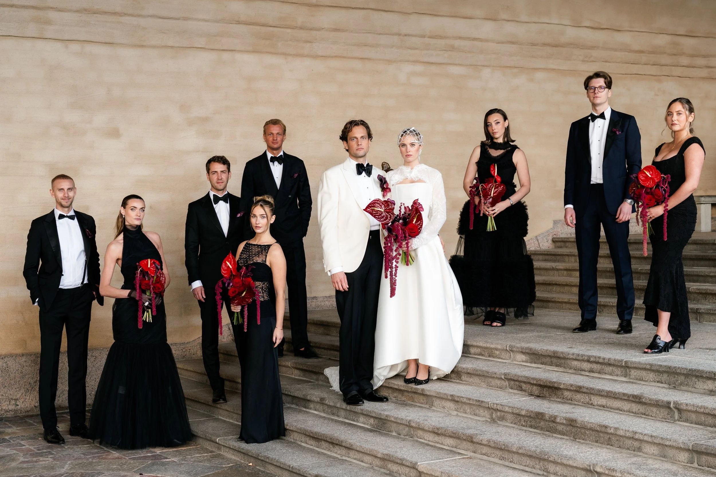 A group of people dressed in formal attire, including tuxedos and evening dresses, standing on the steps of a building. The central couple is dressed in wedding attire, with the bride in a white gown holding a bouquet of red flowers, and the groom in