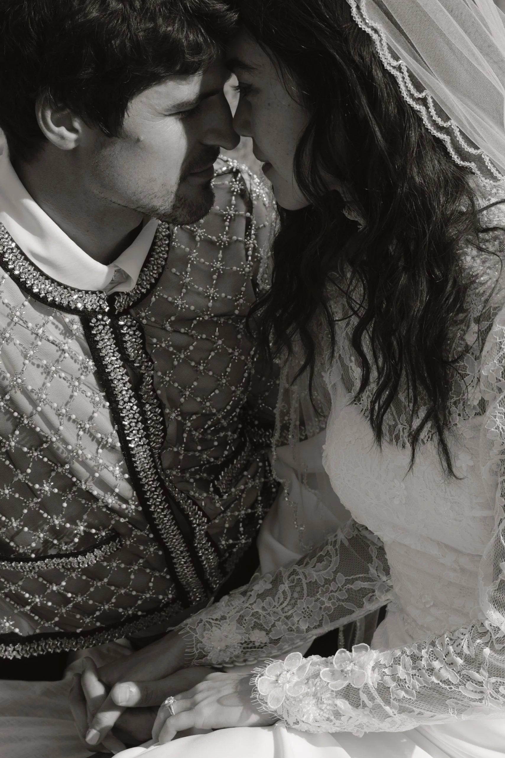 A black-and-white photo of a couple close together, touching foreheads and eyes closed, dressed in wedding attire, holding hands gently.