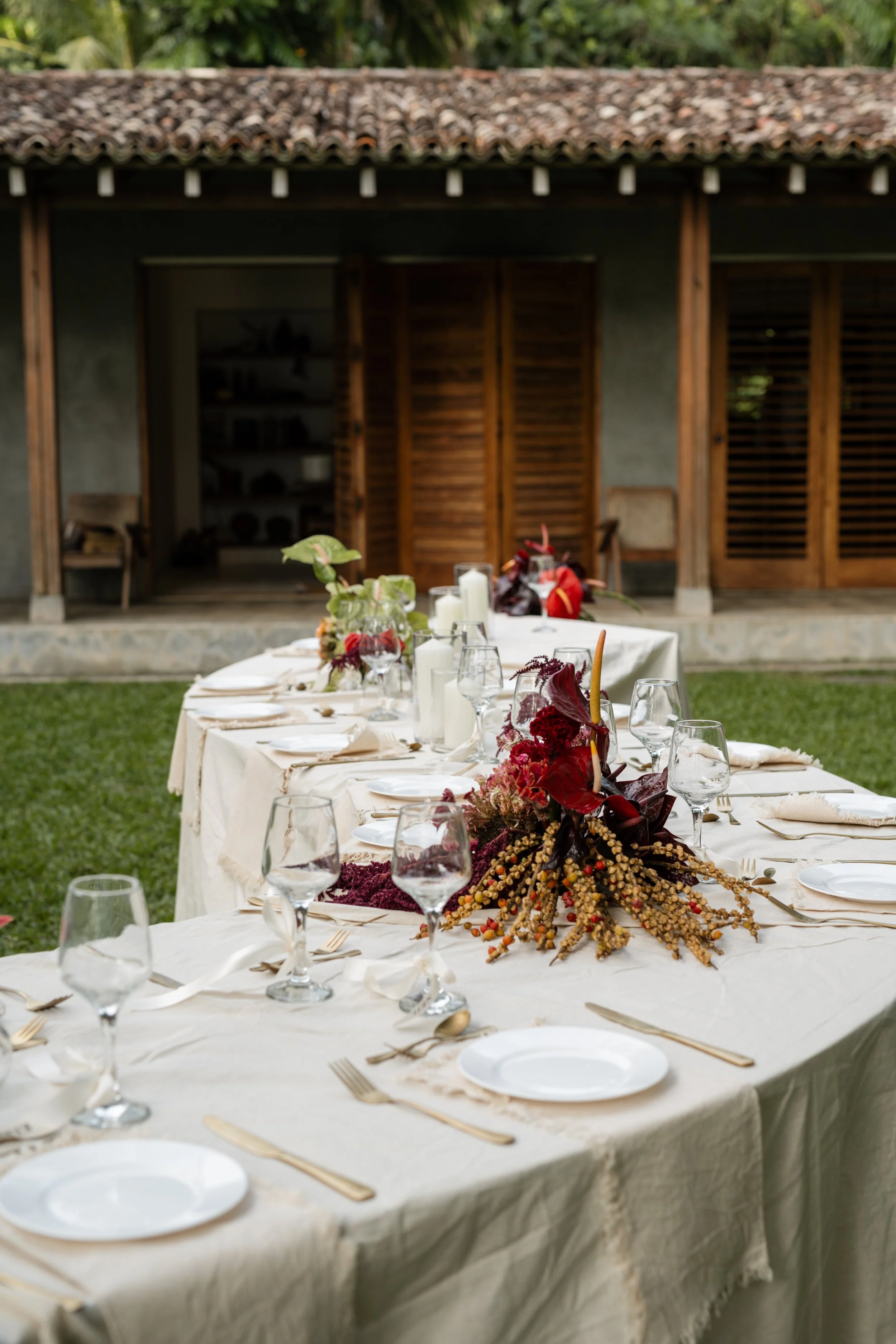 An outdoor dining table decorated with a floral centerpiece and set with plates, silverware, and glasses, in front of a rustic house with open wooden shutters.