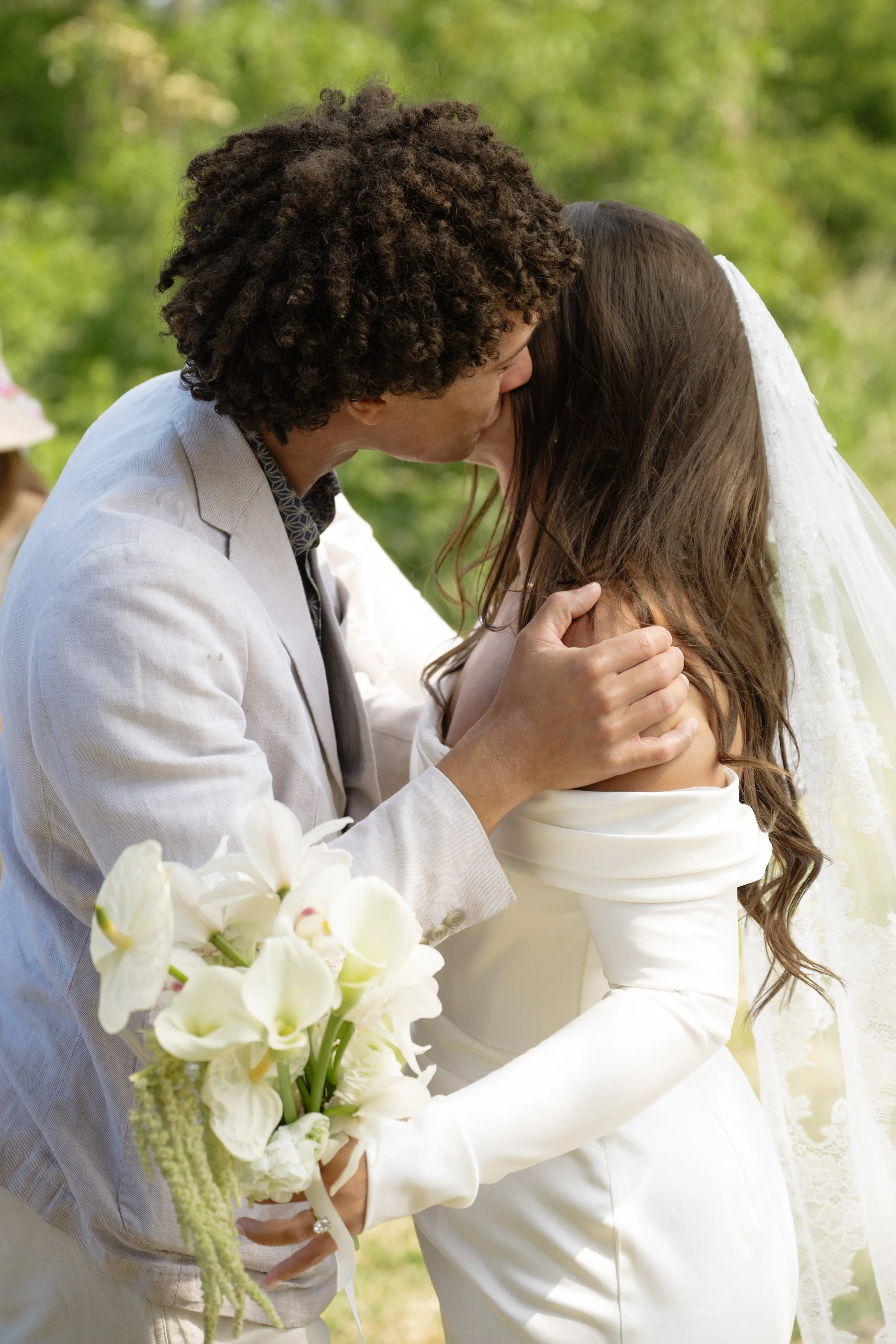 A couple sharing a kiss during their wedding, outdoors surrounded by green trees. The man has curly brown hair and is wearing a light-colored suit. The woman has long brown hair, a white gown with lace details, and is holding a bouquet of white flowe