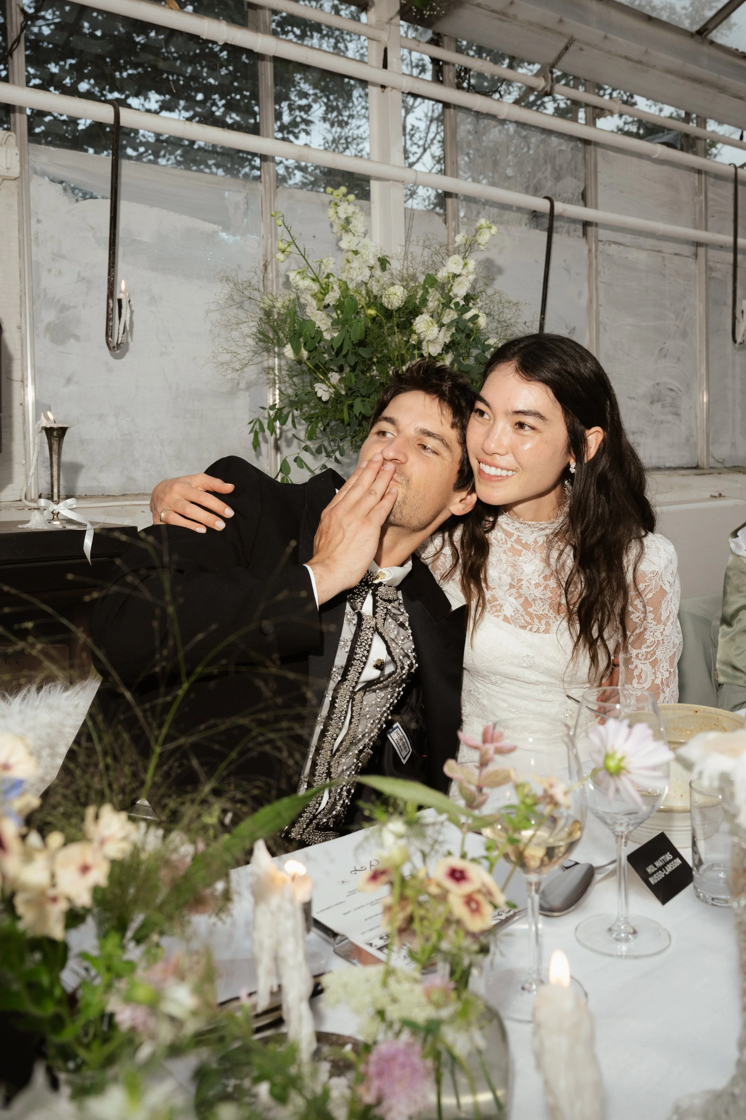 A man and woman at a wedding reception, with the man whispering and the woman smiling, sitting at a decorated table with flowers and candles.