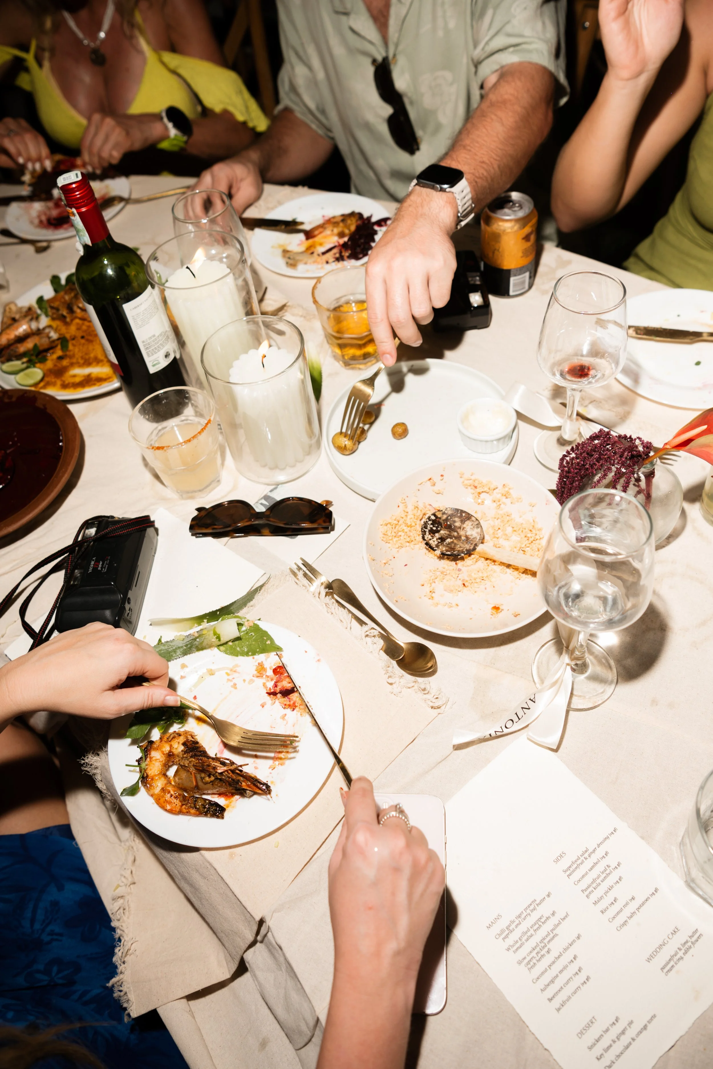 A table with dirty plates, glasses, and various food items at a social gathering, with people dining and drinks present.