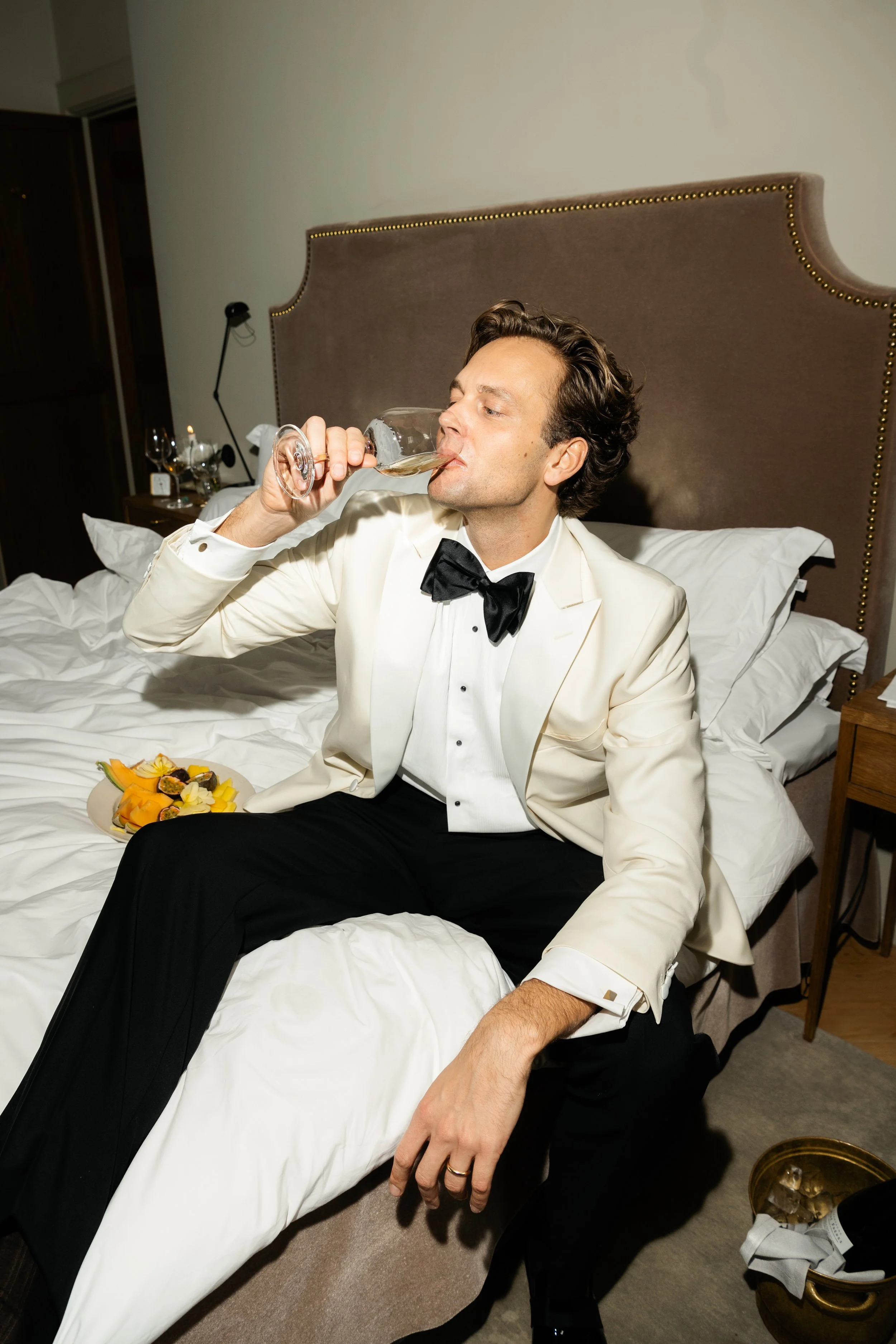 A man in tuxedo with a white jacket and black bow tie, sitting on a bed, drinking wine from a glass, with a plate of food nearby.