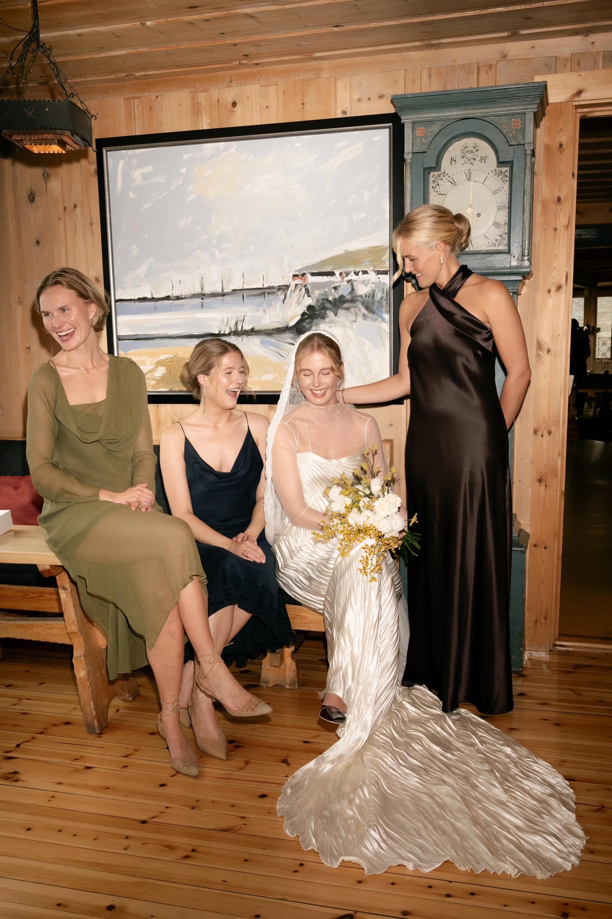 A bride and bridesmaids sharing a joyful moment indoors. The bride is sitting with a bouquet, wearing a champagne-colored satin wedding dress with a veil. Three women are gathered around, smiling and engaging with her. The setting has wooden walls, a
