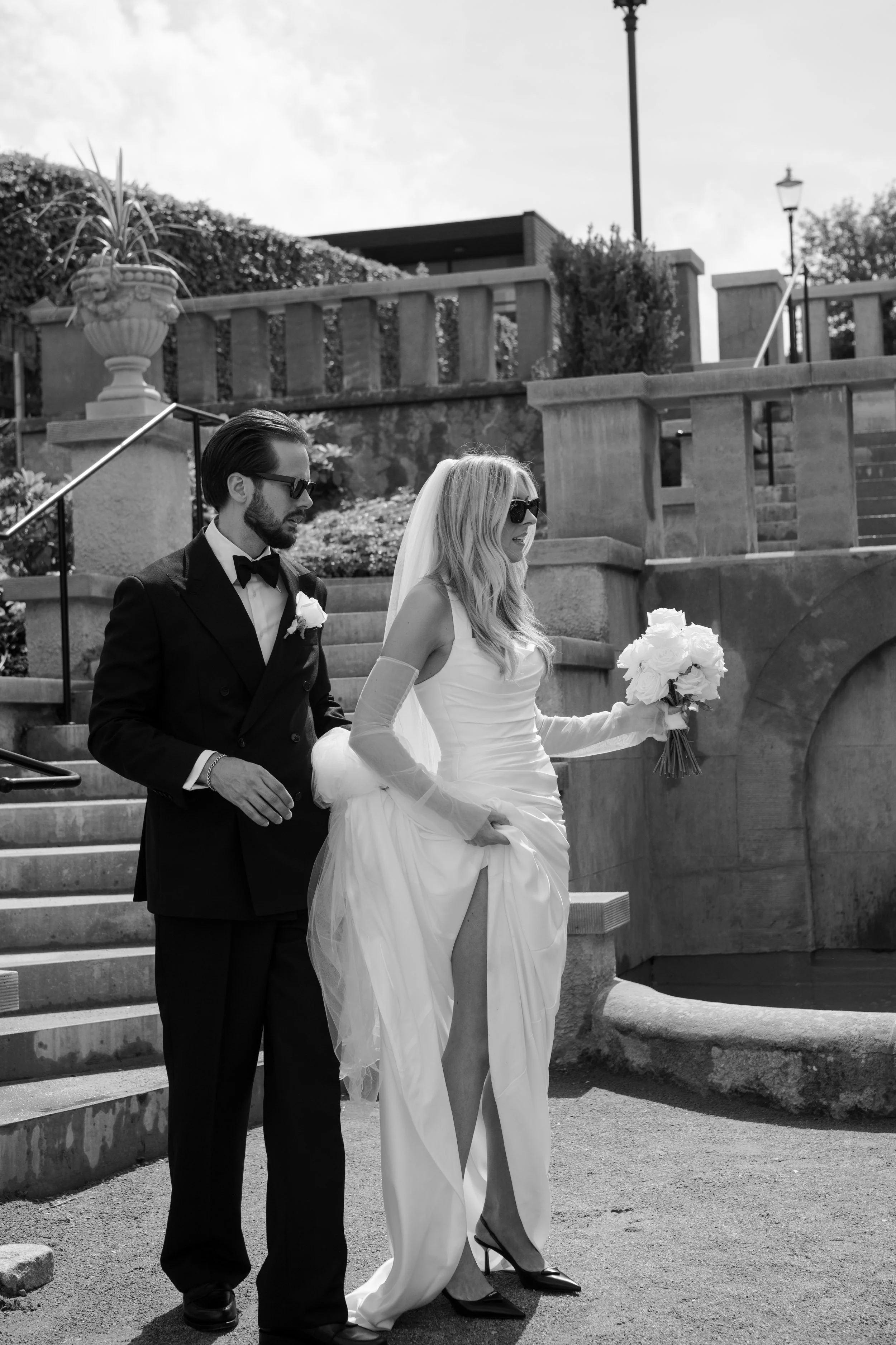 A bride in a wedding dress holding a bouquet and a groom in a tuxedo walking down stairs outdoors.