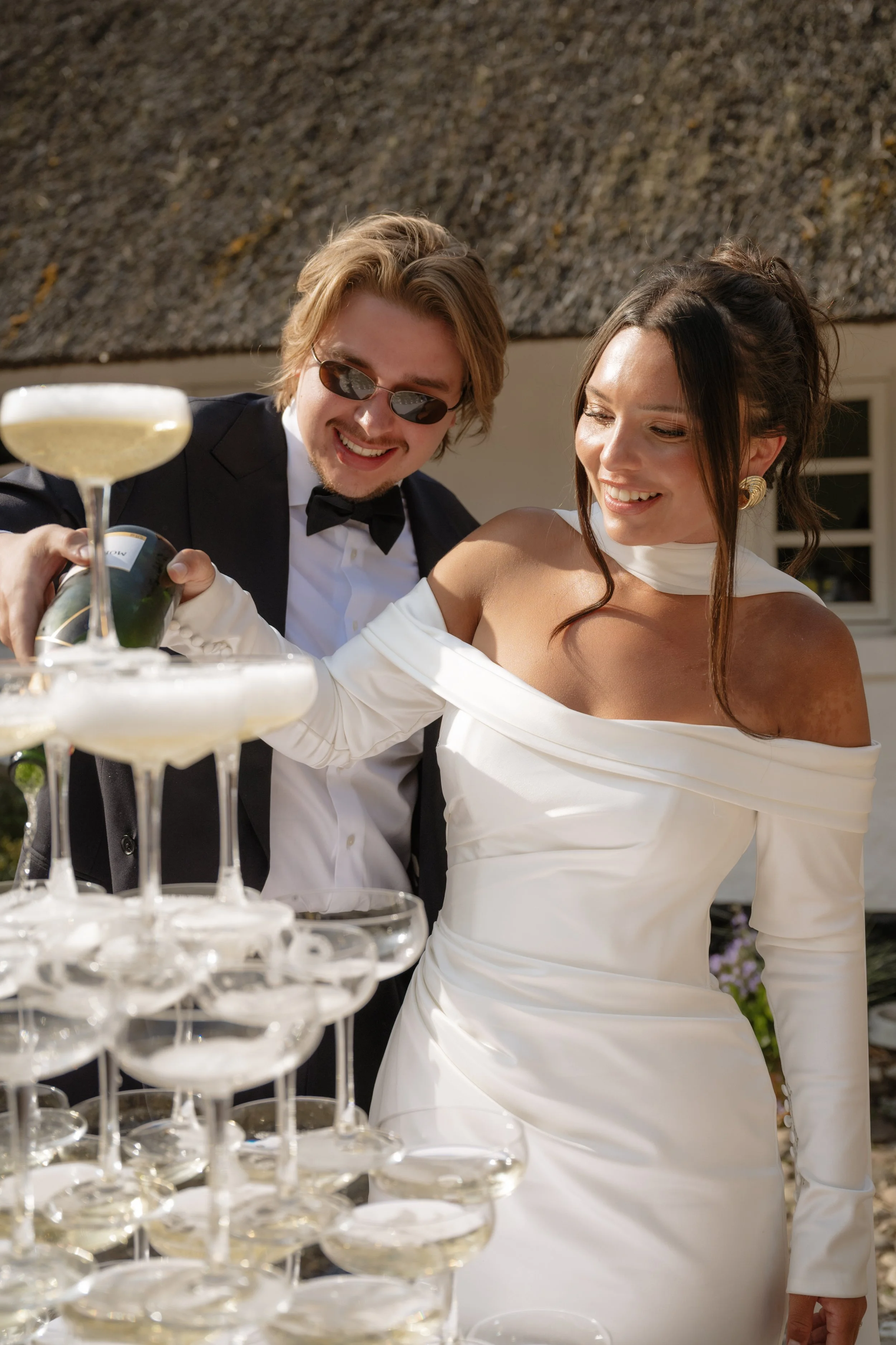 A bride and groom celebrating at a wedding reception with a champagne tower, the groom pouring champagne into the top glass.