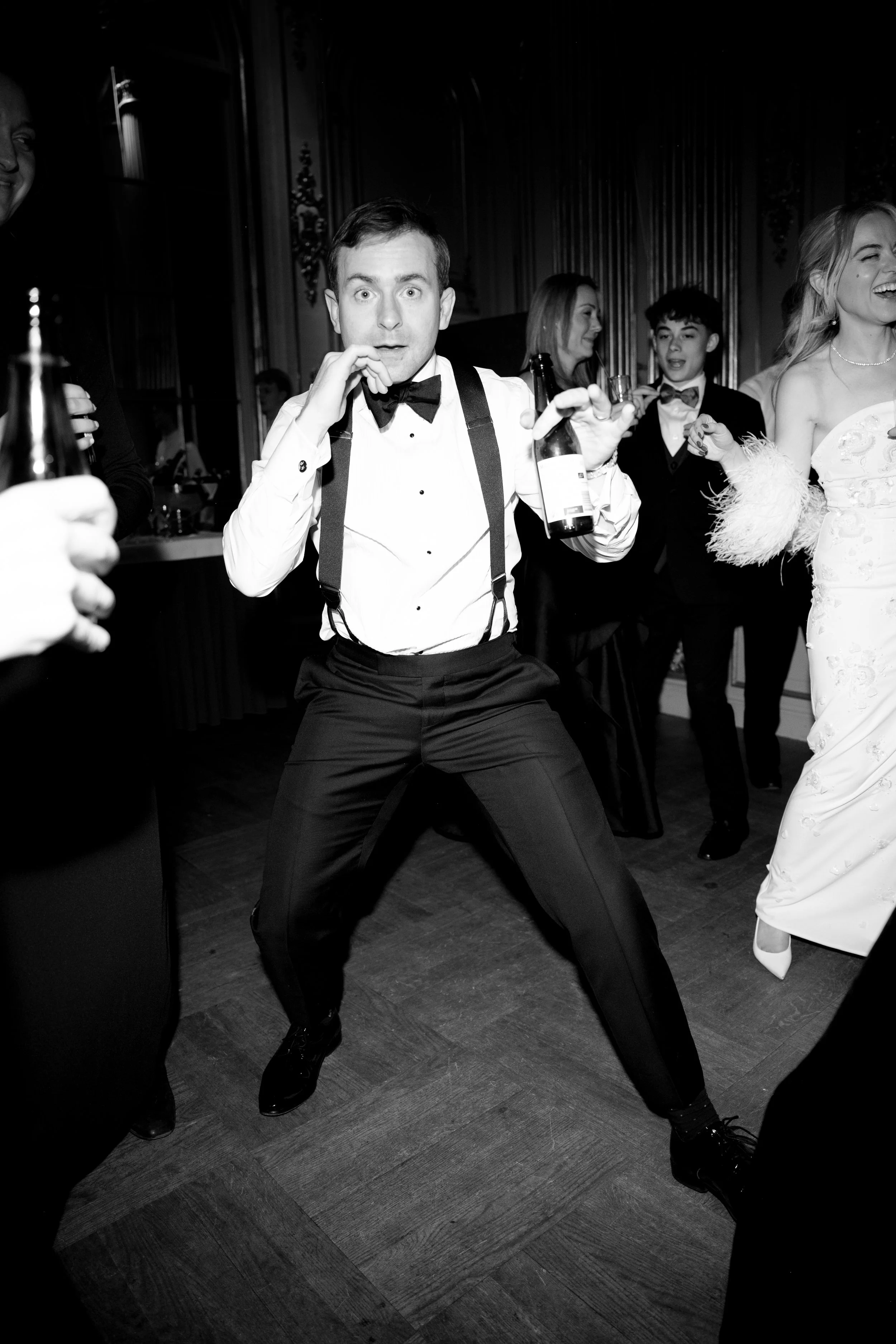 Black and white photo of a man in formal attire dancing at a party, holding a beer bottle, with other guests in the background enjoying the celebration.