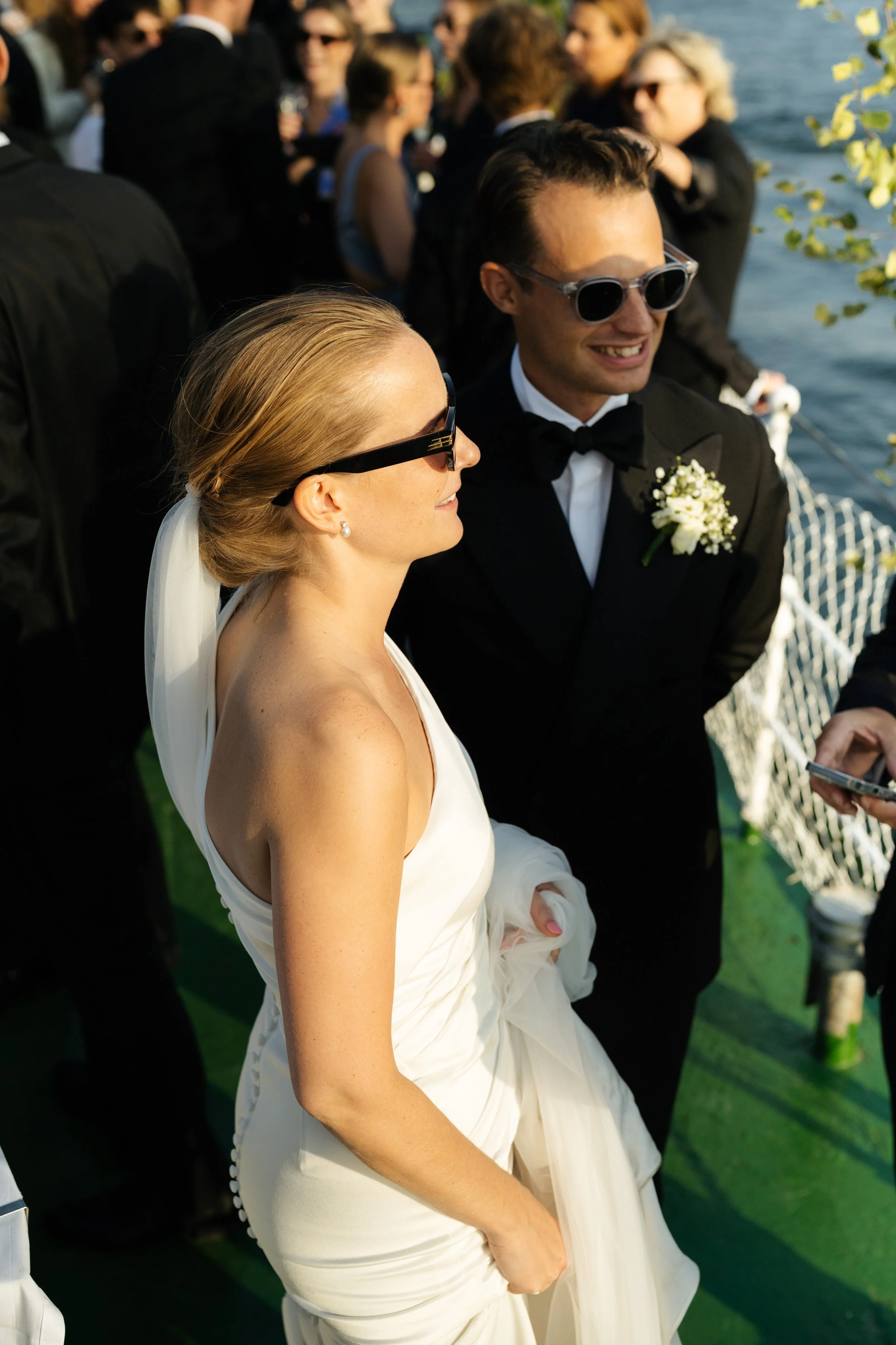 A bride and groom wearing sunglasses, smiling, at their wedding by a lake, with guests in the background.