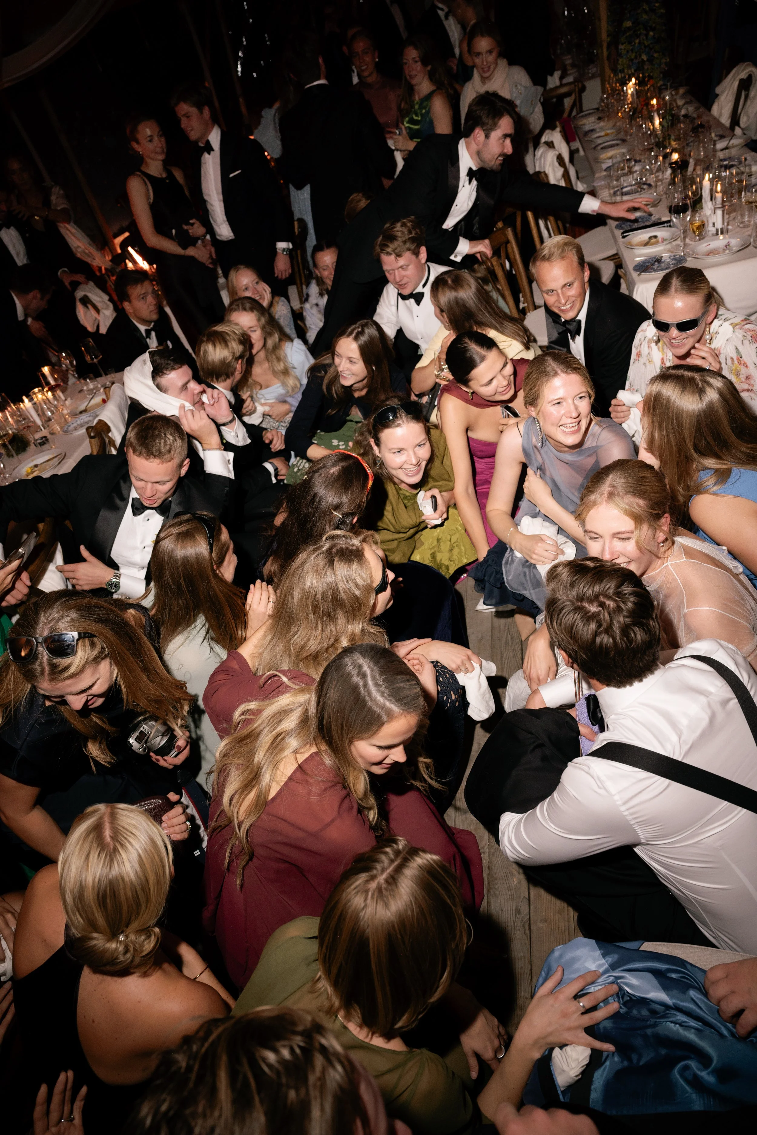 A large group of people at a formal party or wedding reception, sitting and standing around a crowded dance floor, smiling and enjoying the celebration.