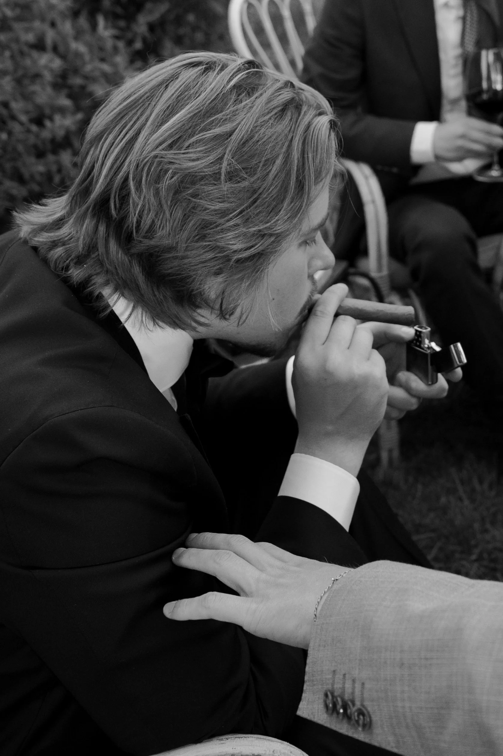 A young man with shoulder-length hair smoking a cigar while seated at a formal event, wearing a dark suit, with other guests in the background.