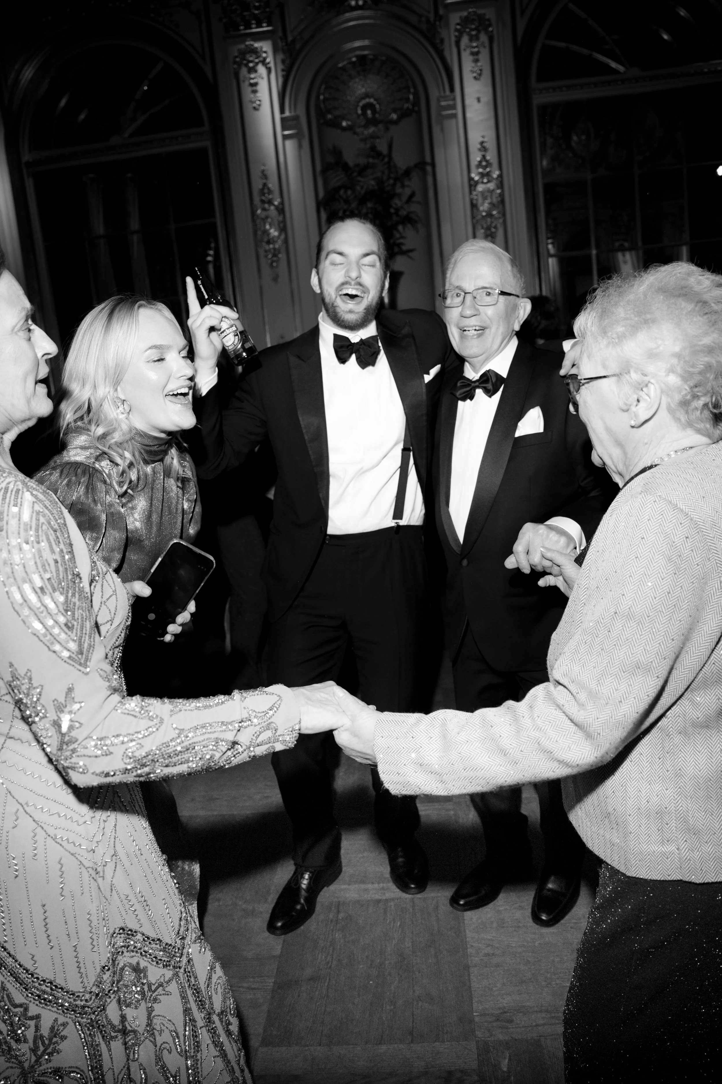 People dancing and celebrating at a formal event, with a man holding a drink and others smiling and holding hands.