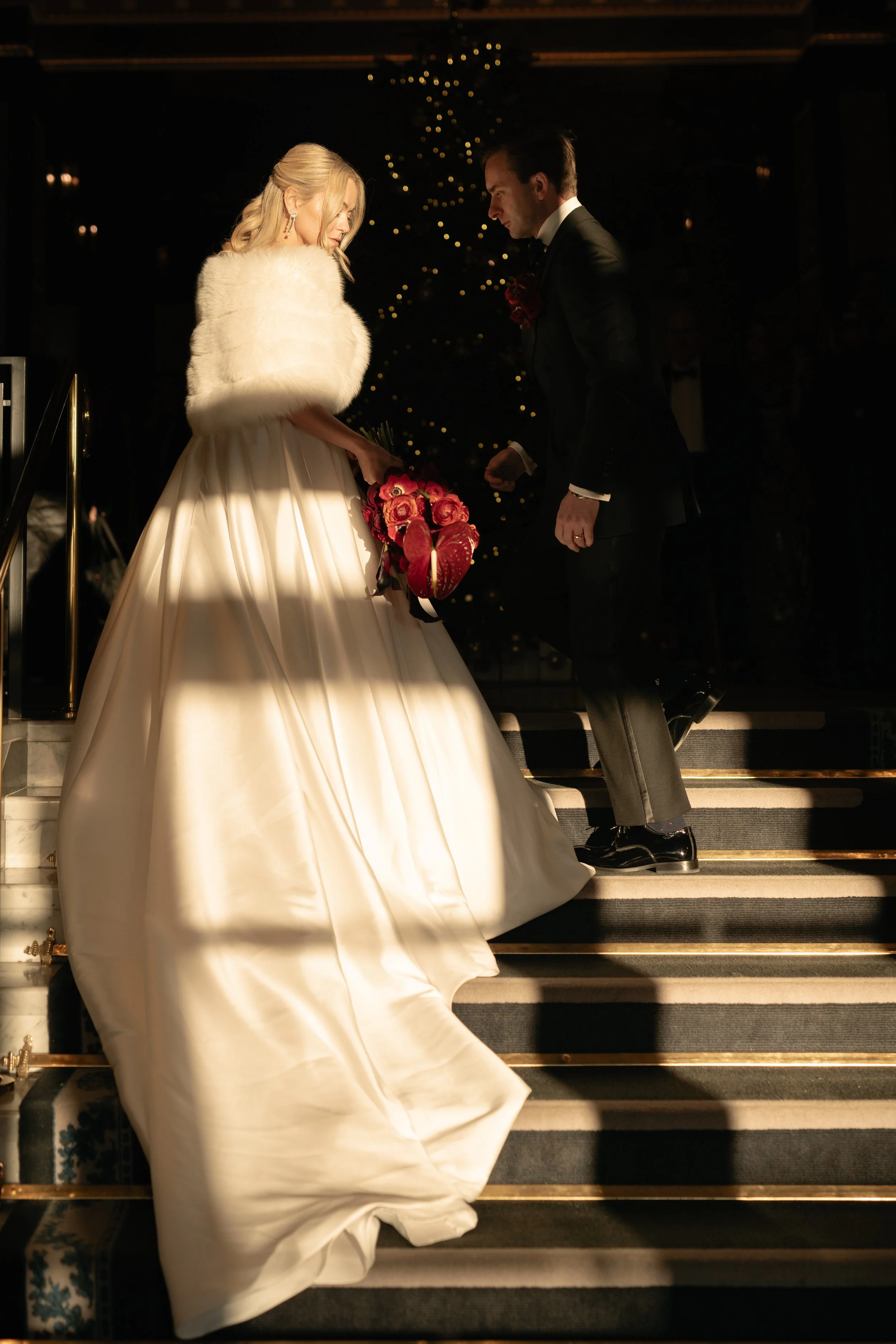 A bride in a white gown holding a bouquet of pink roses, standing on stairs with a groom in a black tuxedo proposing.