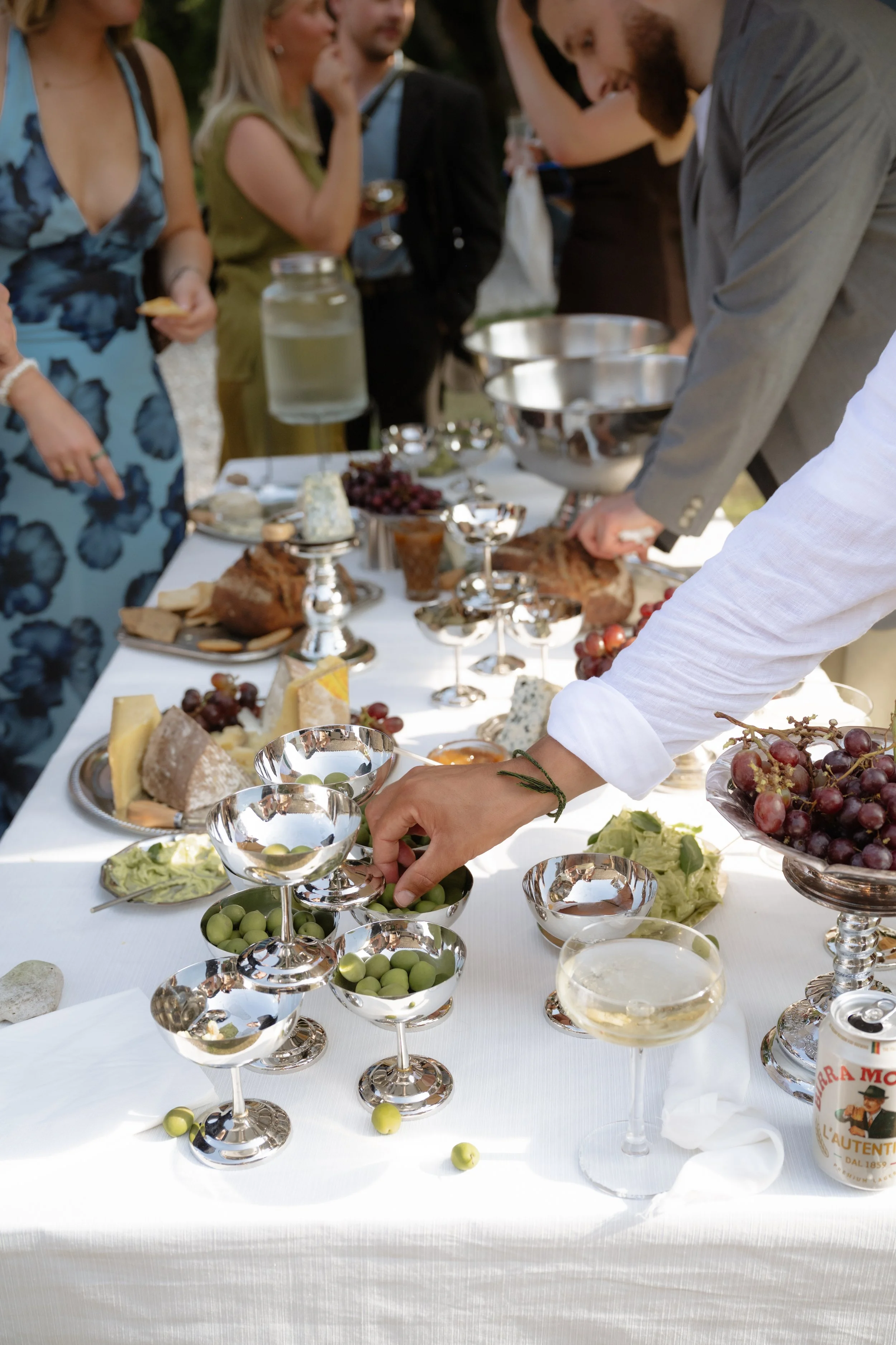 People gather around a table with cheese, grapes, and glasses, serving themselves at a social outdoor event or celebration.