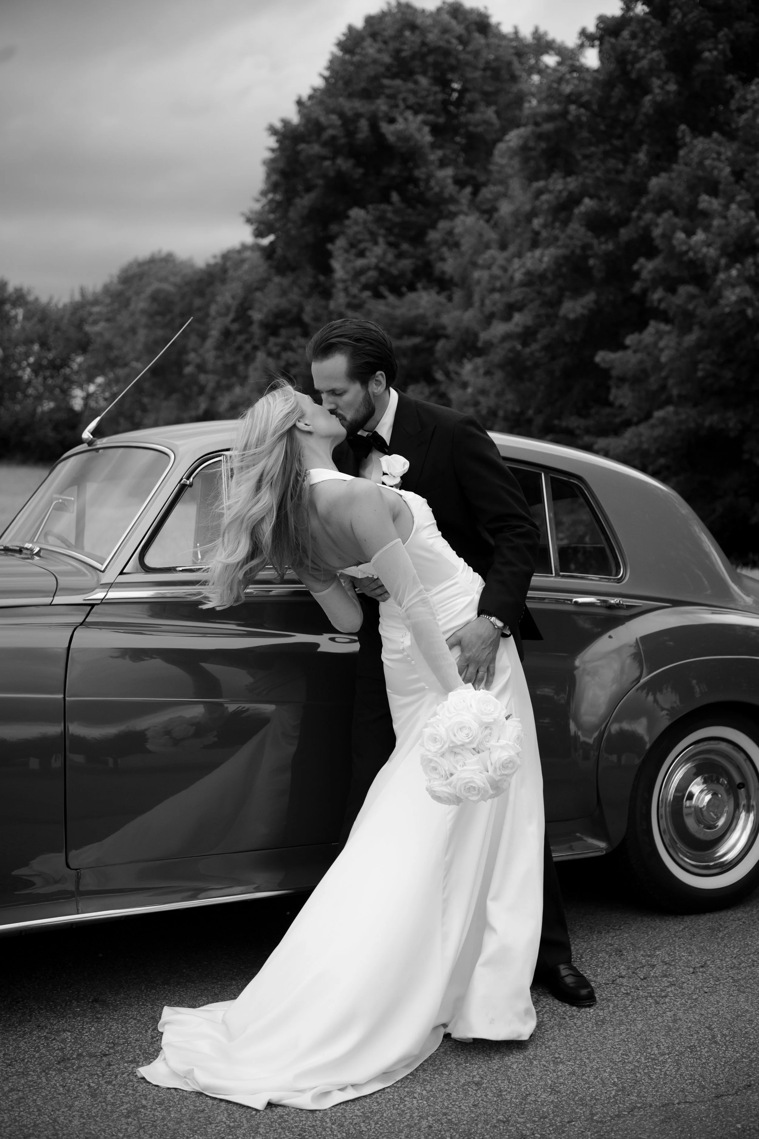 Black and white photo of a bride and groom kissing in front of a vintage car, with trees in the background.