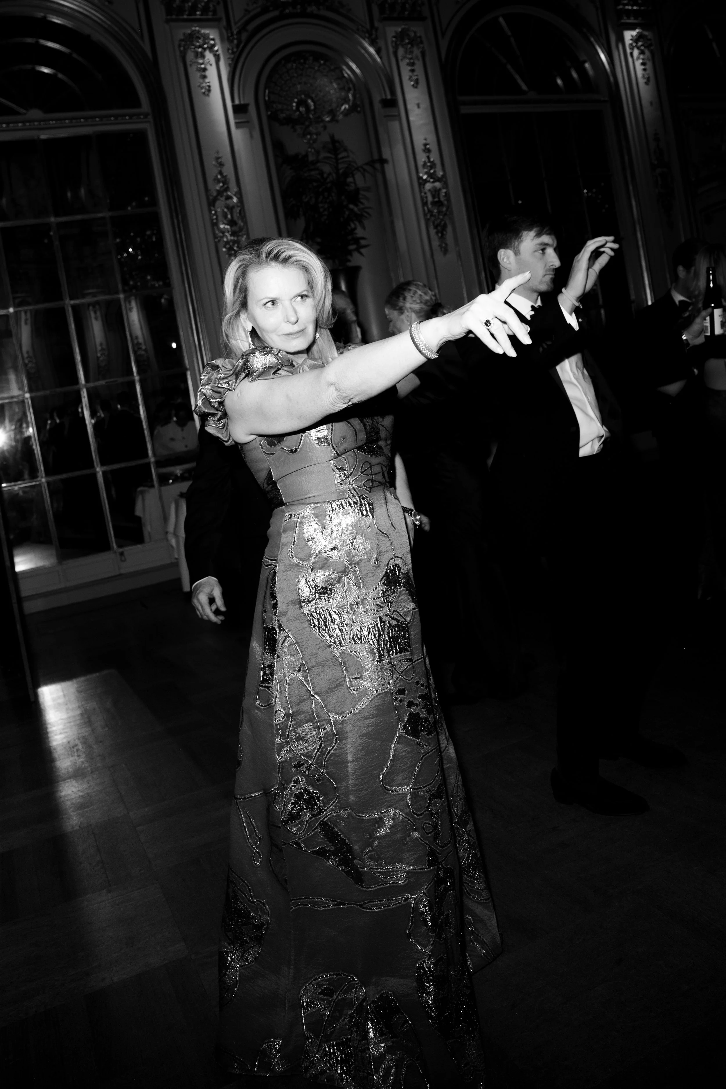 A woman in an elegant dress appears to be dancing or posing at a formal event in a decorated ballroom with large windows and ornate wall details, black and white photo.