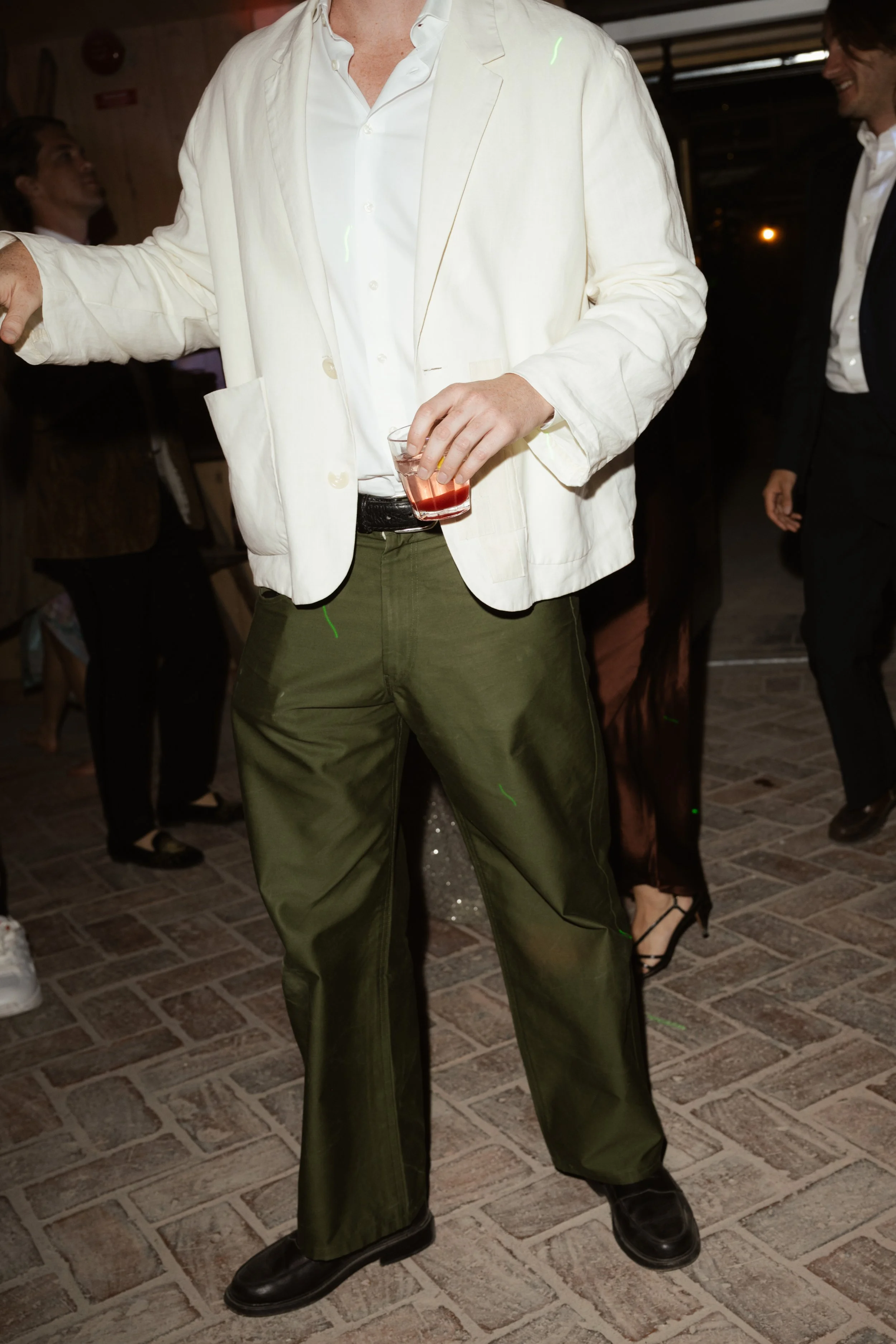 Man dressed in a white blazer, white shirt, and green pants holding a drink at a social gathering.