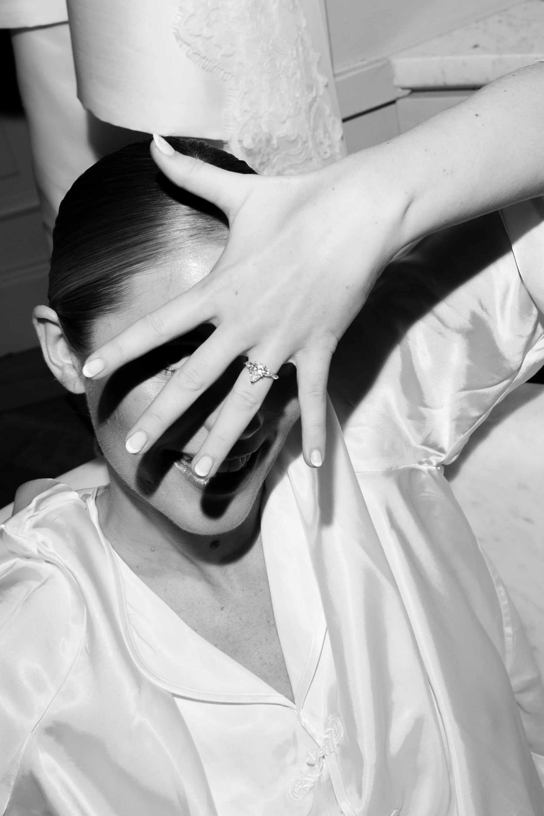 A woman with slicked-back hair and wearing a satin robe is covering her face with her hand, which has rings, while smiling.