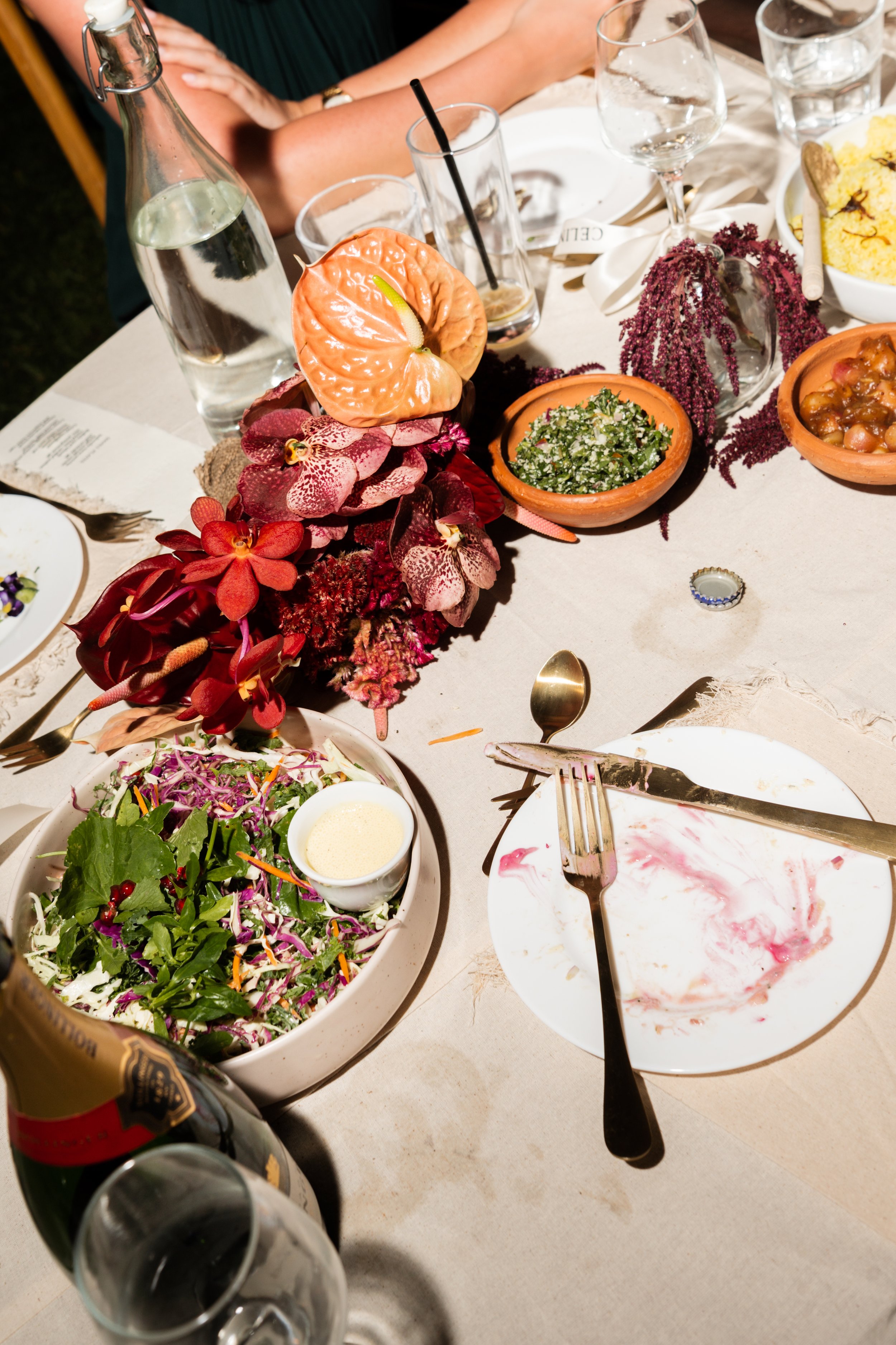 A dinner table with scattered dishes, glasses, and utensils. Centerpiece of vibrant flowers, a bottle of water, and various plates of salad and side dishes, some partially finished.