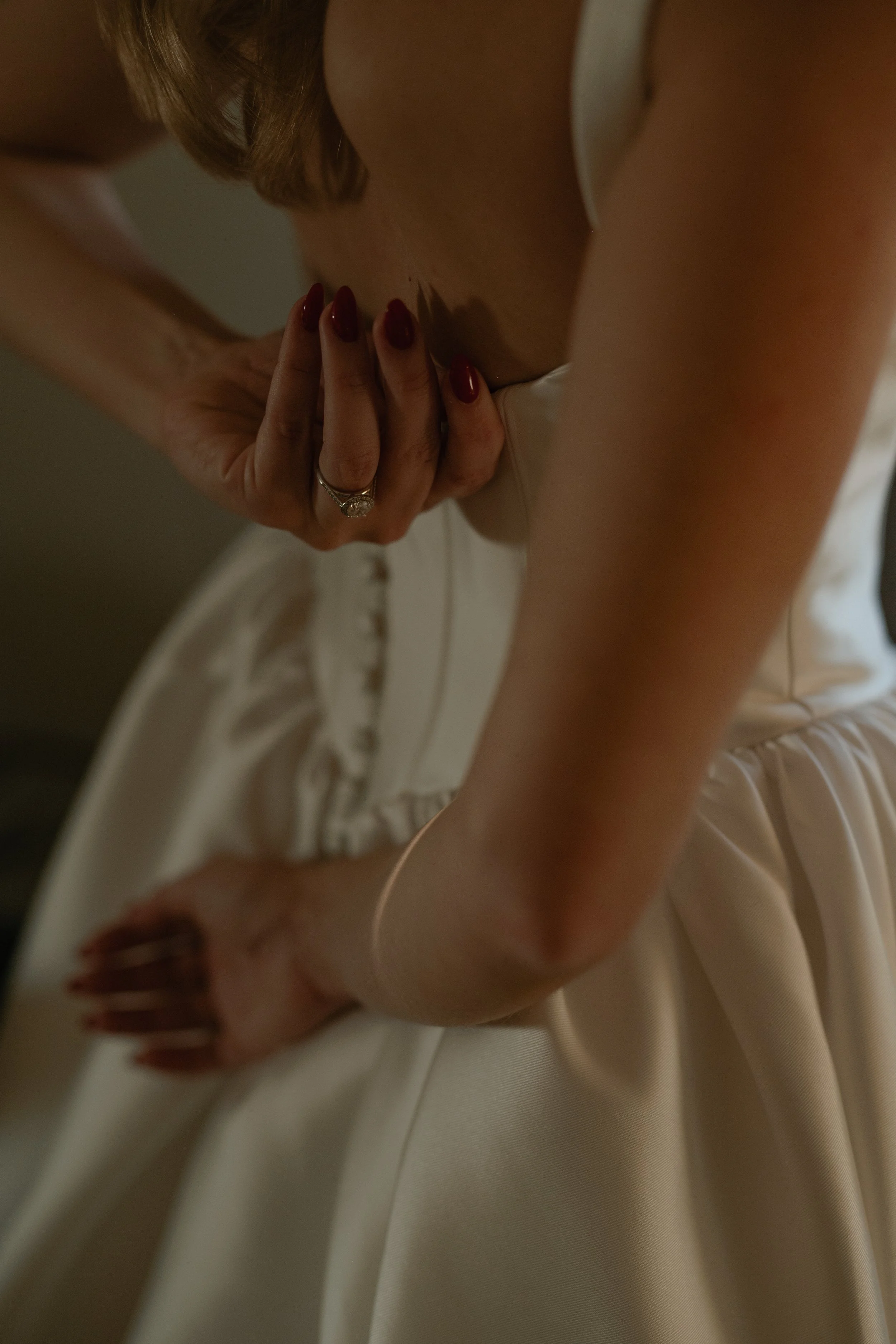 A woman in a white dress with red painted nails and a diamond ring on her finger is holding her chest with one hand, showing her engagement ring, while the other hand rests on her hip.