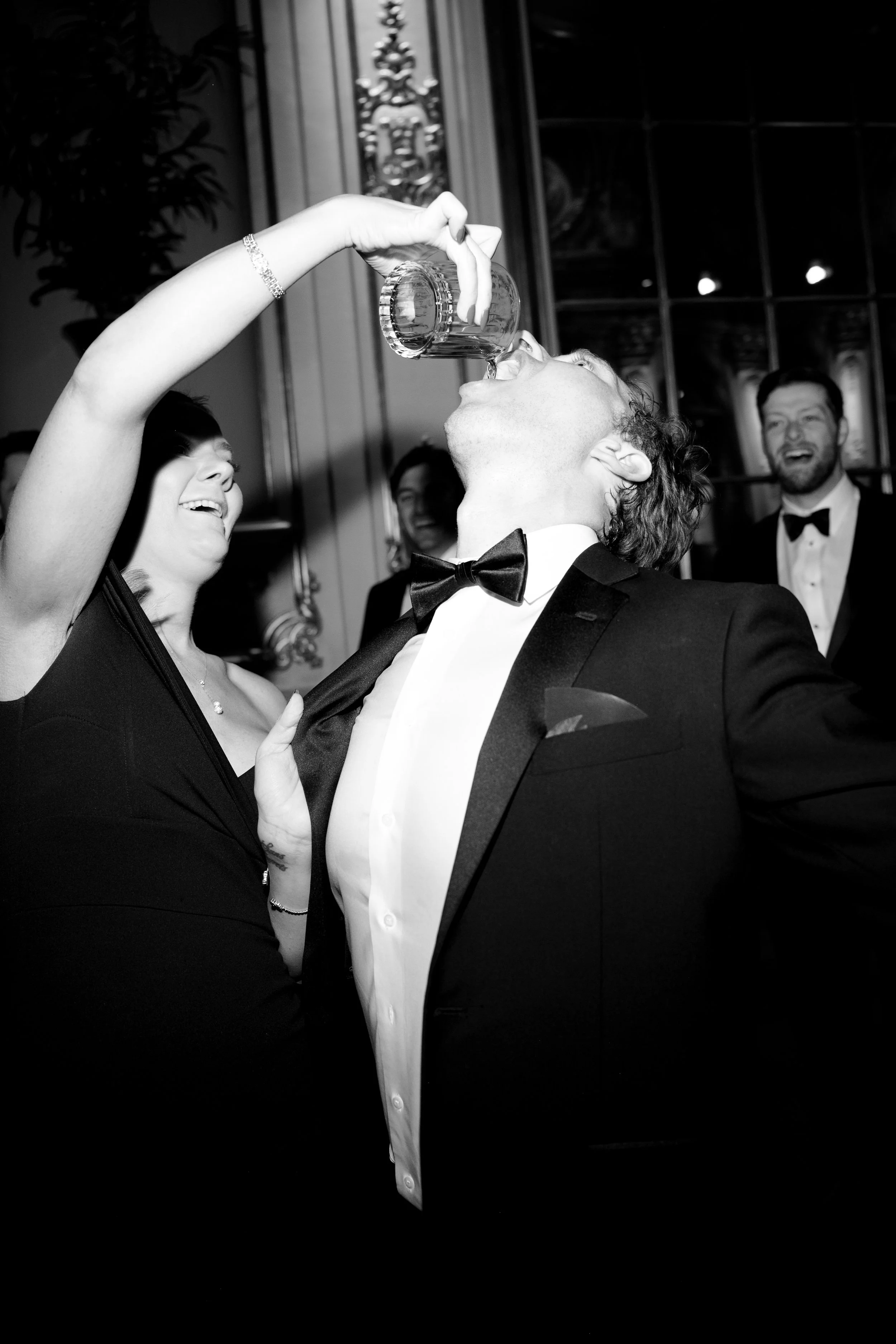 People celebrating at a formal event, with one man in a tuxedo taking a shot from a glass held by a woman in a black dress, others smiling in the background.