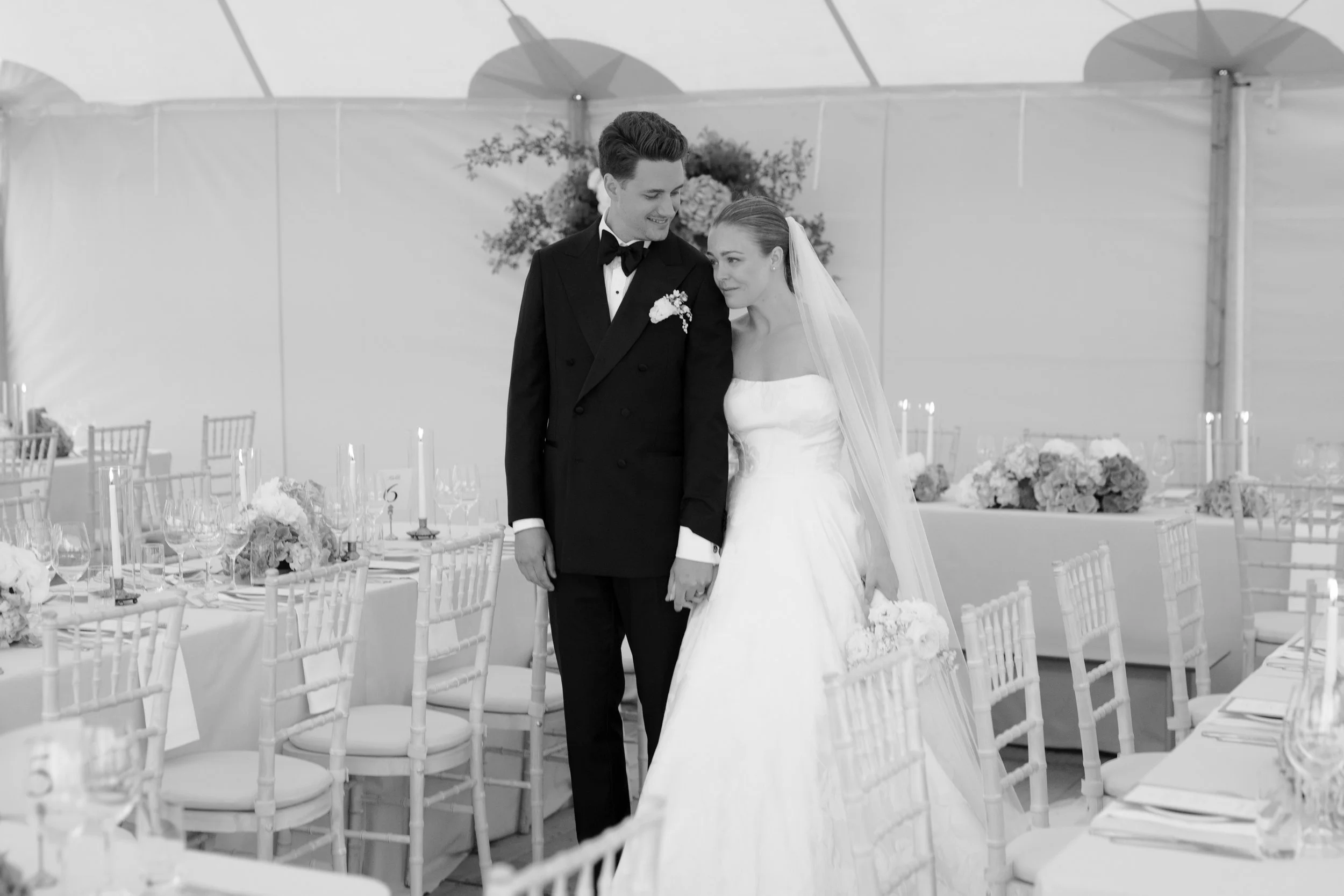 Black and white photo of a bride and groom holding hands at their wedding reception, standing in a decorated tent with tables and chairs.