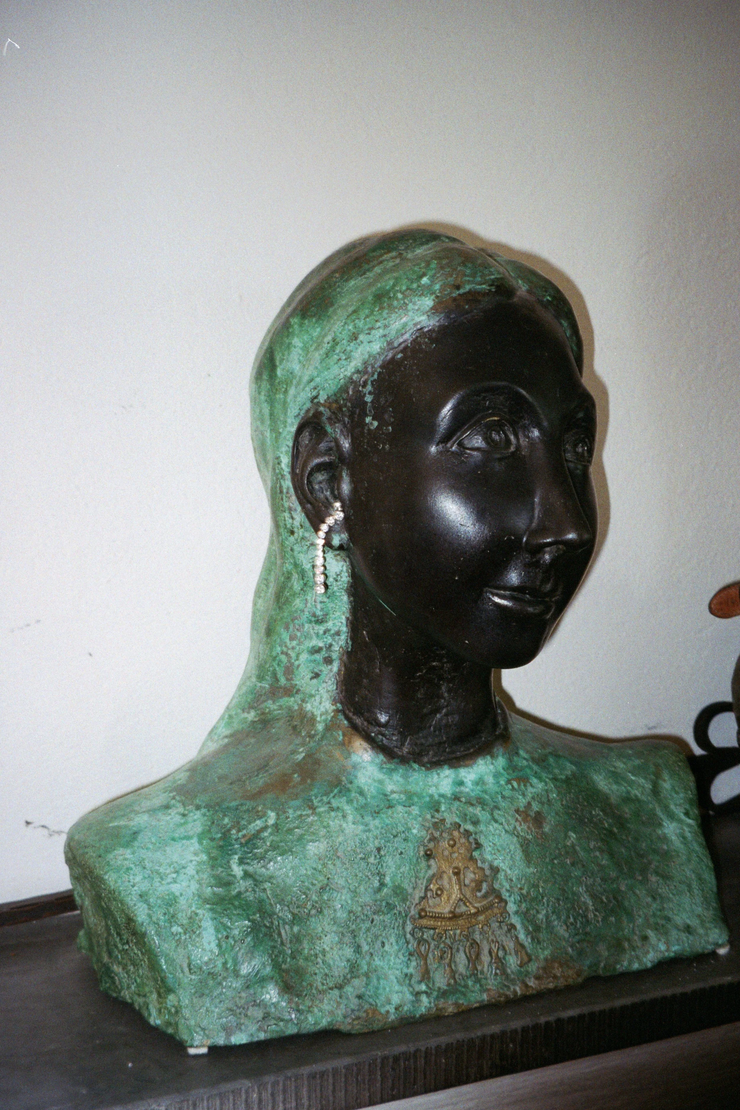 A bust sculpture of a woman with a dark face and greenish-green textured attire, wearing a pearl earring and a detailed necklace. She has a serene expression.