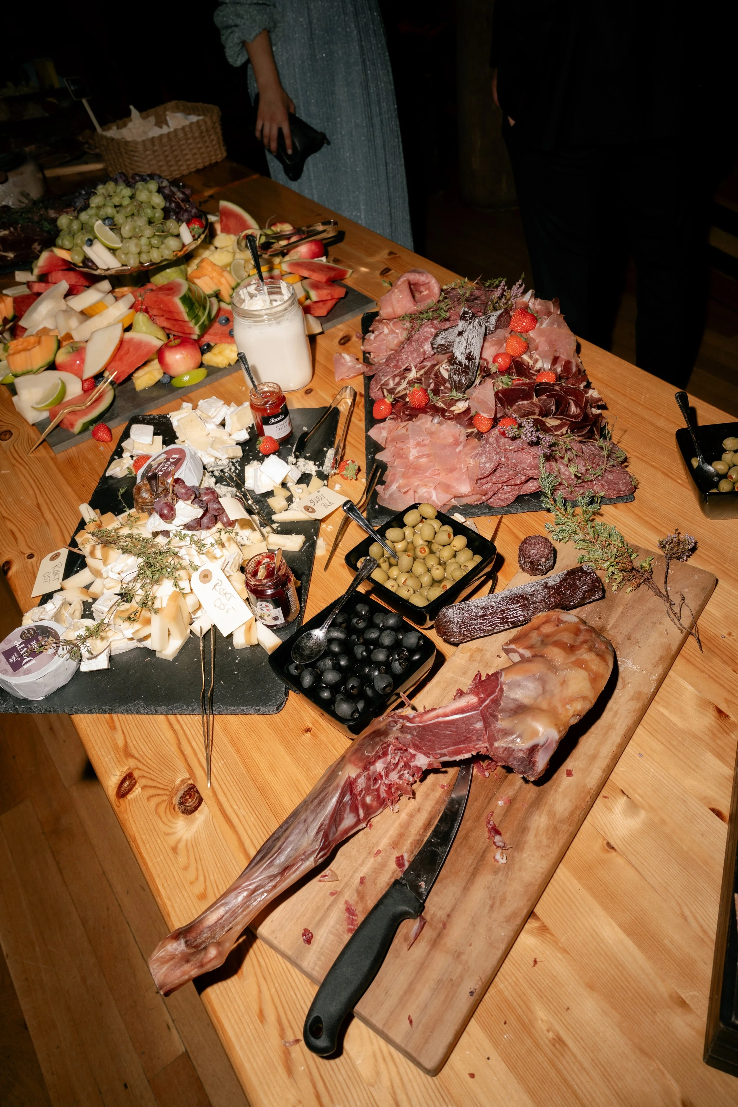 A cheese and charcuterie display with various cheeses, cured meats, olives, and fruits on a wooden table. There's a large leg of prosciutto or ham with a knife stuck in it, alongside jars of jam and small bowls of pickles and olives.