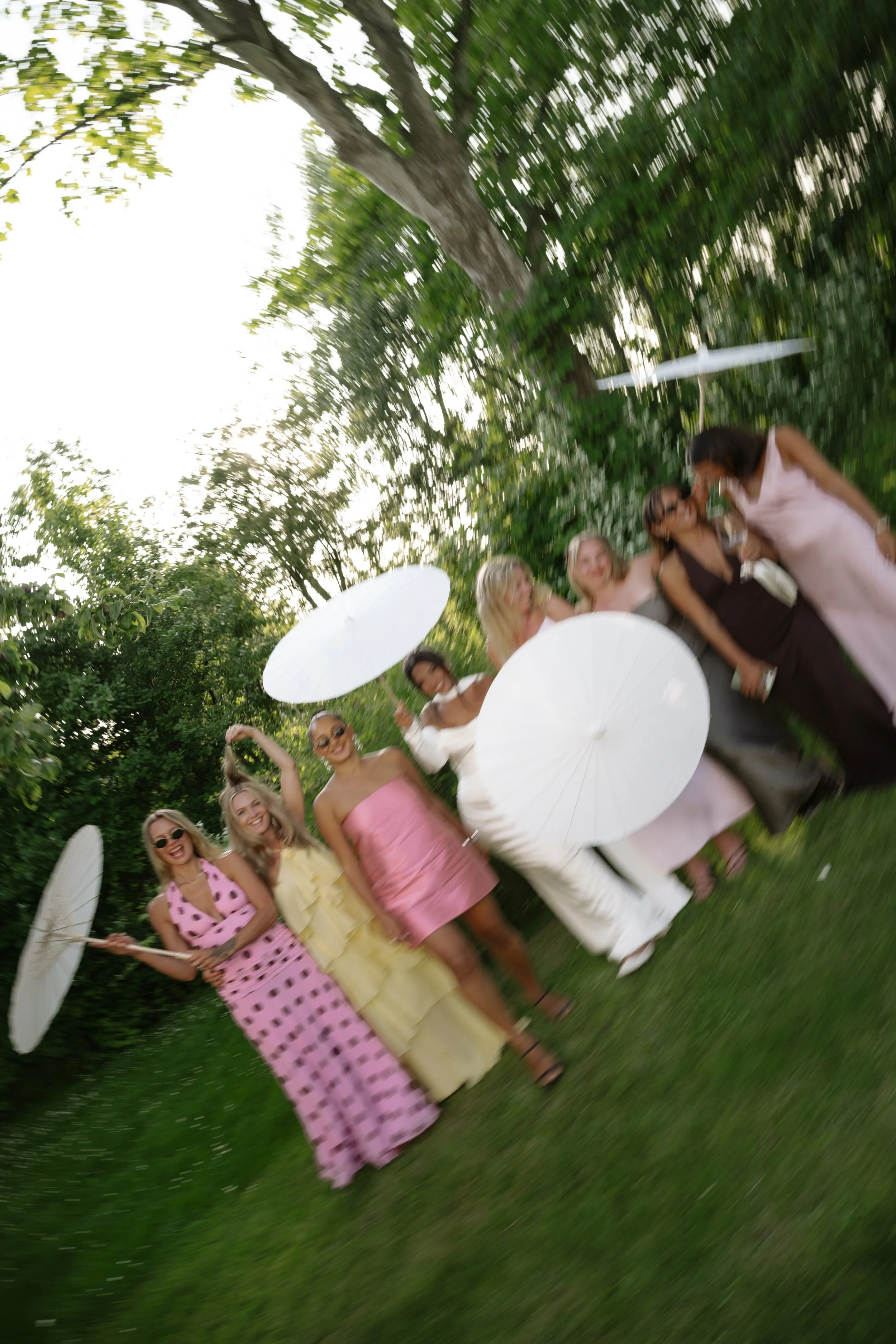 A group of women in colorful summer dresses holding white umbrellas, standing on grass in a lush green outdoor setting, smiling and enjoying a sunny day.