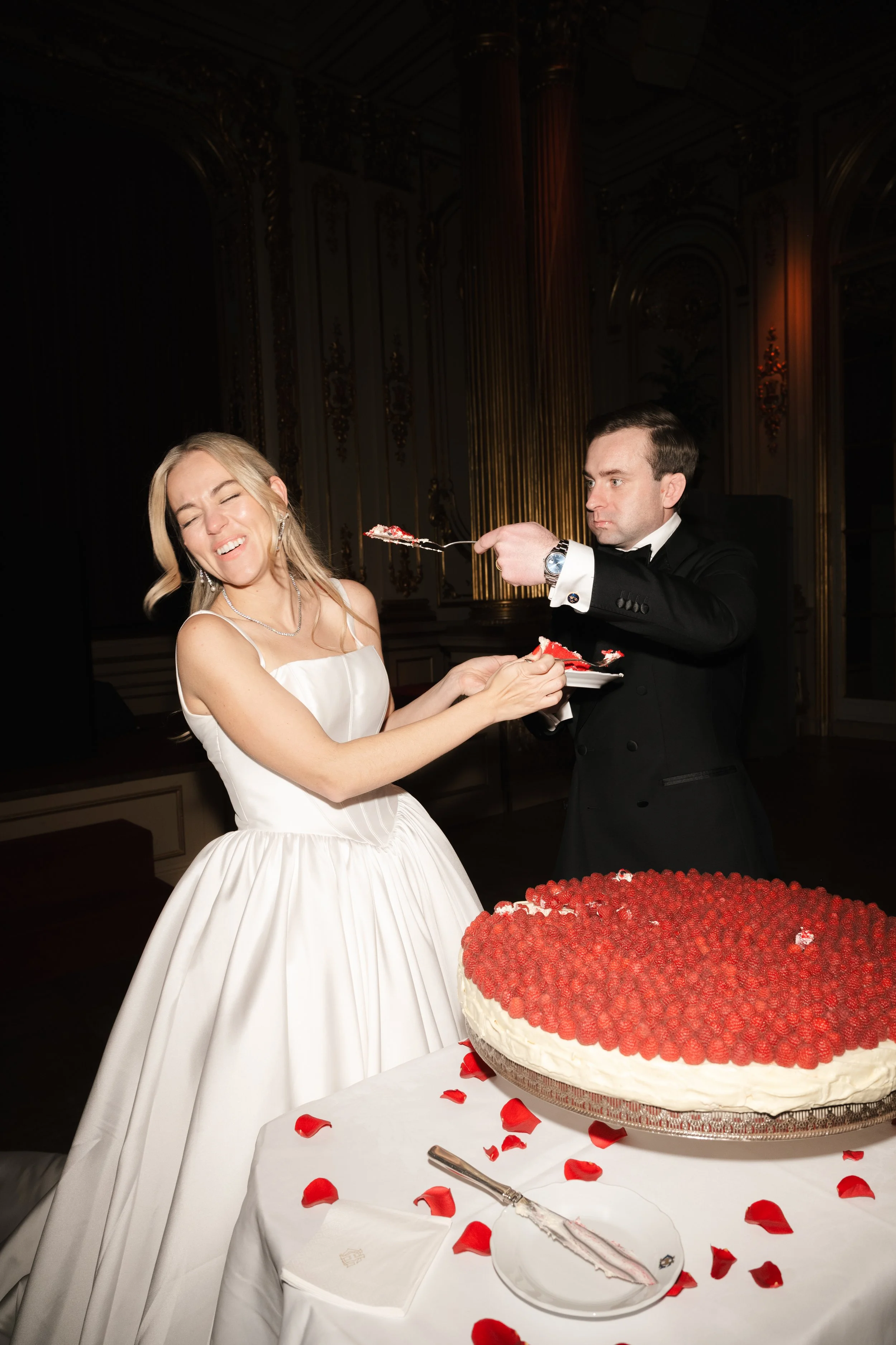 Bride in white wedding gown smiling as groom in black tuxedo serves her a piece of wedding cake at a wedding reception.