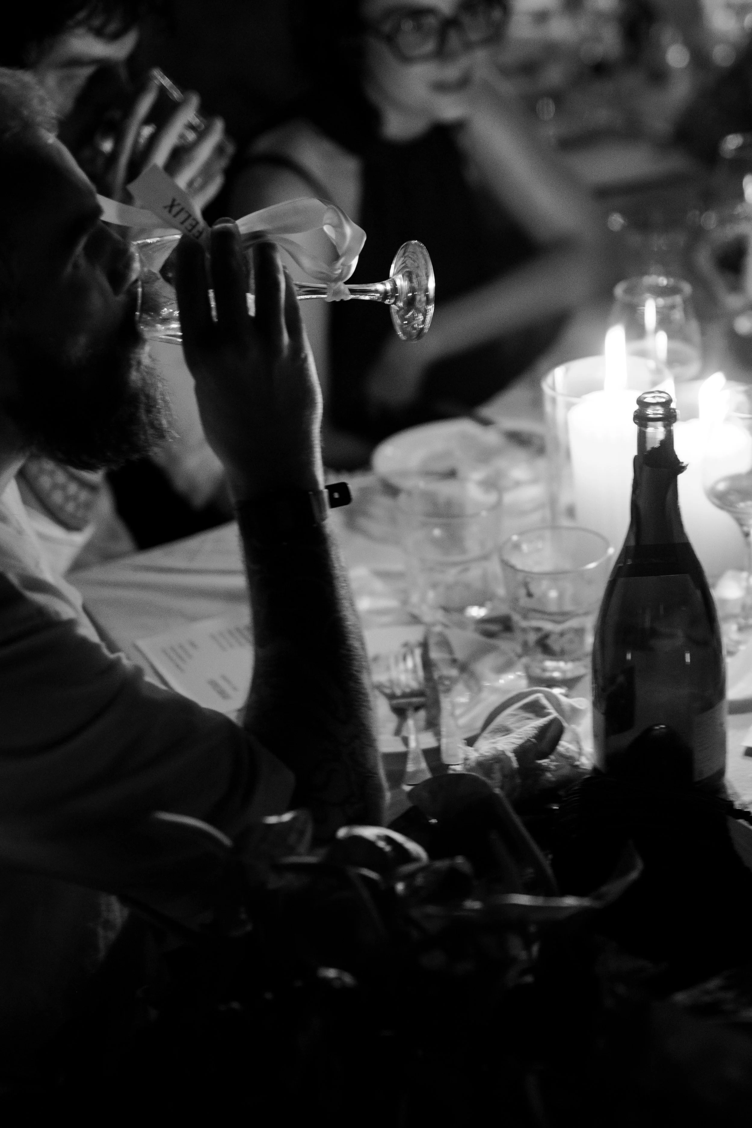 A black and white photo of a man drinking from a glass at a dinner table, with a woman sitting nearby, various glasses, a bottle, and lit candles.