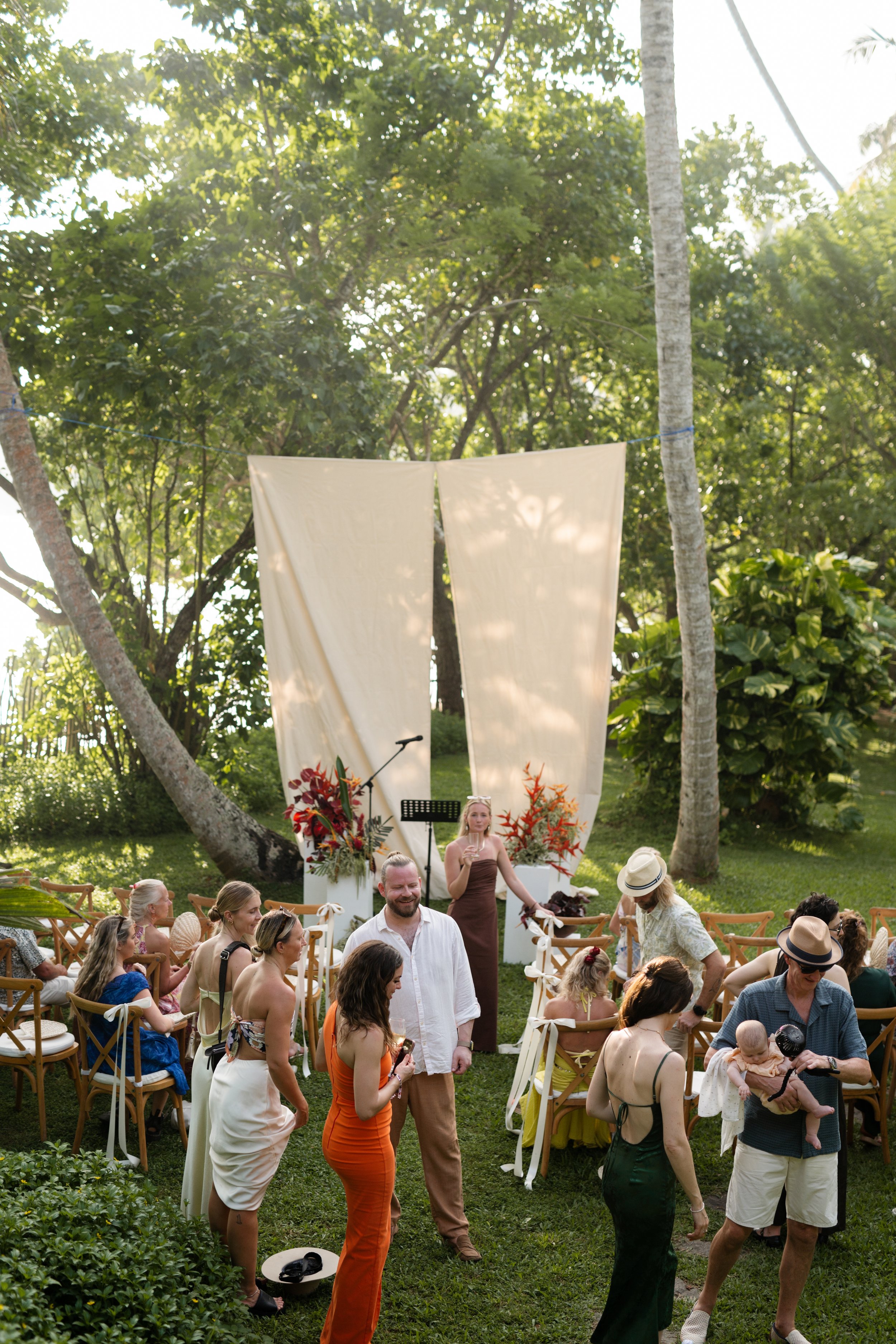 People gathered outdoors for a wedding or celebration with a natural backdrop of trees and grass. There are chairs, floral arrangements, and a person speaking at a microphone on a stage.