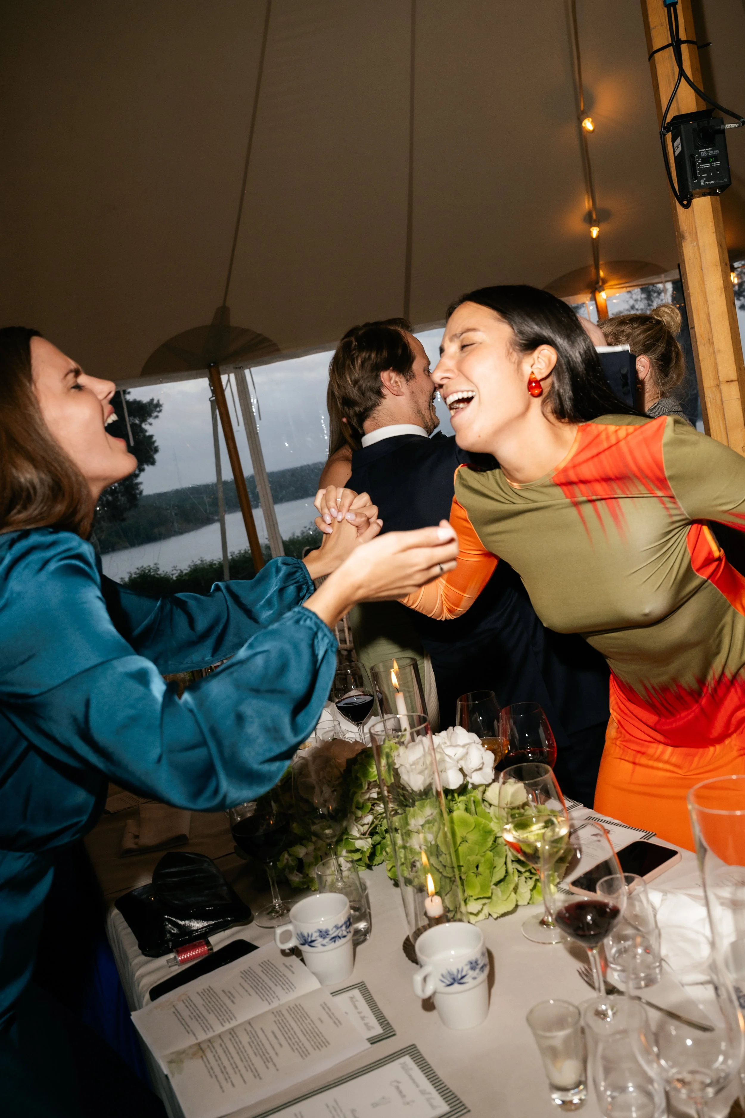 People celebrating at a dinner party, smiling, laughing, and dancing together in a festive setting with drinks and floral centerpieces.