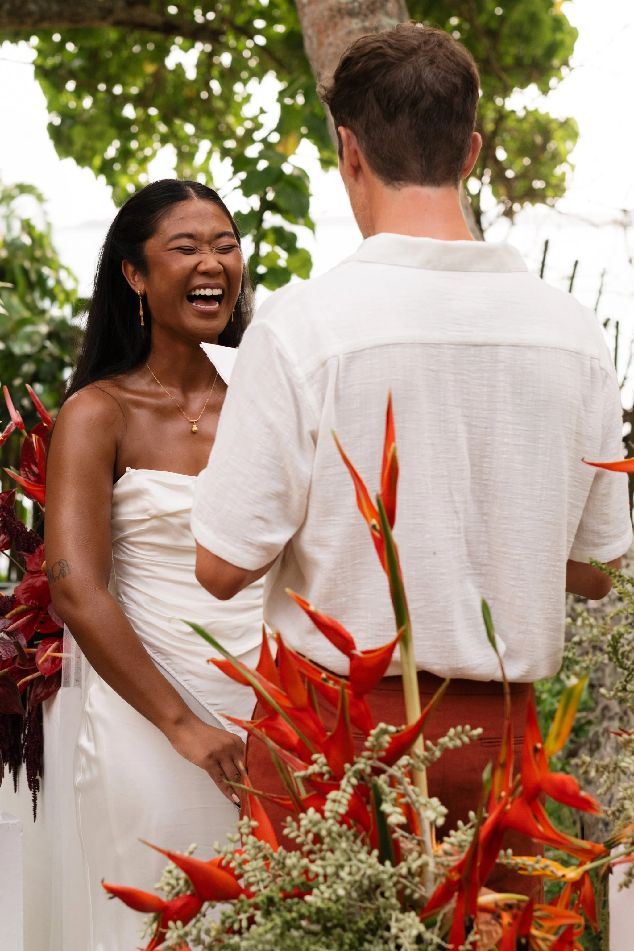 A woman with long black hair and a white strapless dress, laughing and smiling, stands opposite a man with short brown hair and a white shirt, during an outdoor event surrounded by colorful flowers and green foliage.