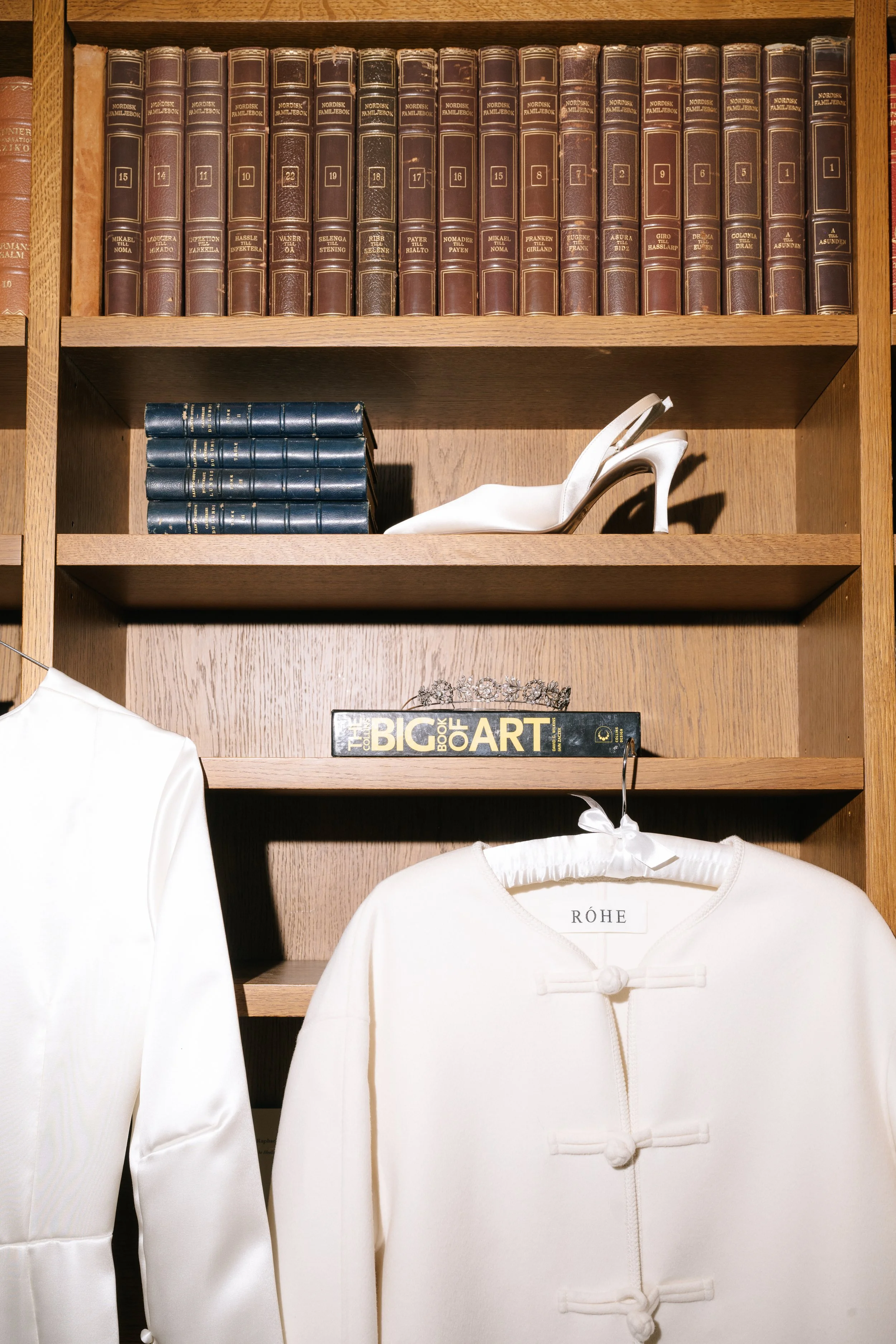 A wooden bookshelf with classic brown leather-bound books on the top shelf, a stack of blue hardcover books and a white high-heeled shoe on the middle shelf, a book titled the big book of art and a tiara on the lower shelf, and two white garments han