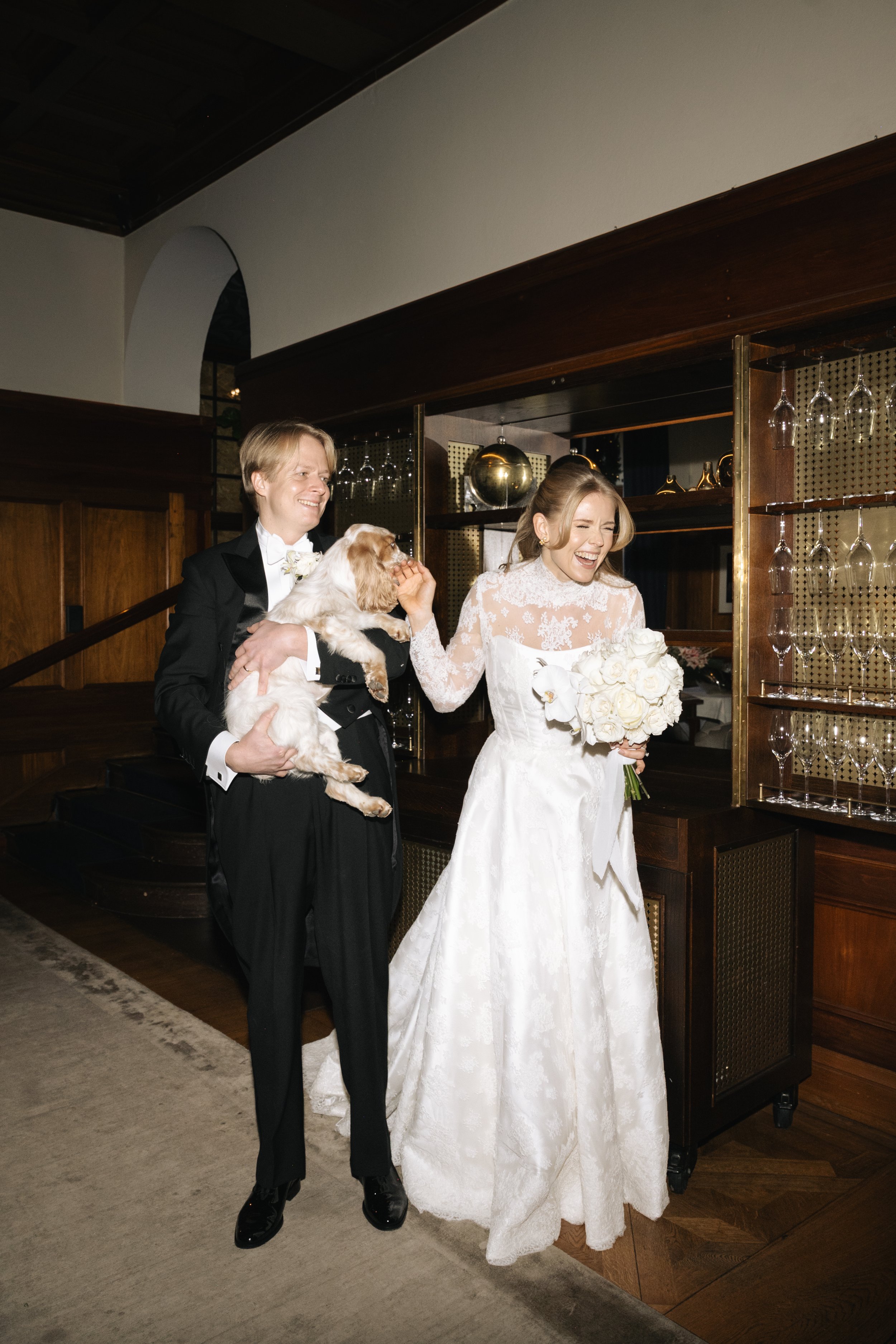 A couple in wedding attire, the groom holding a dog, standing close and smiling, in a well-decorated indoor setting.