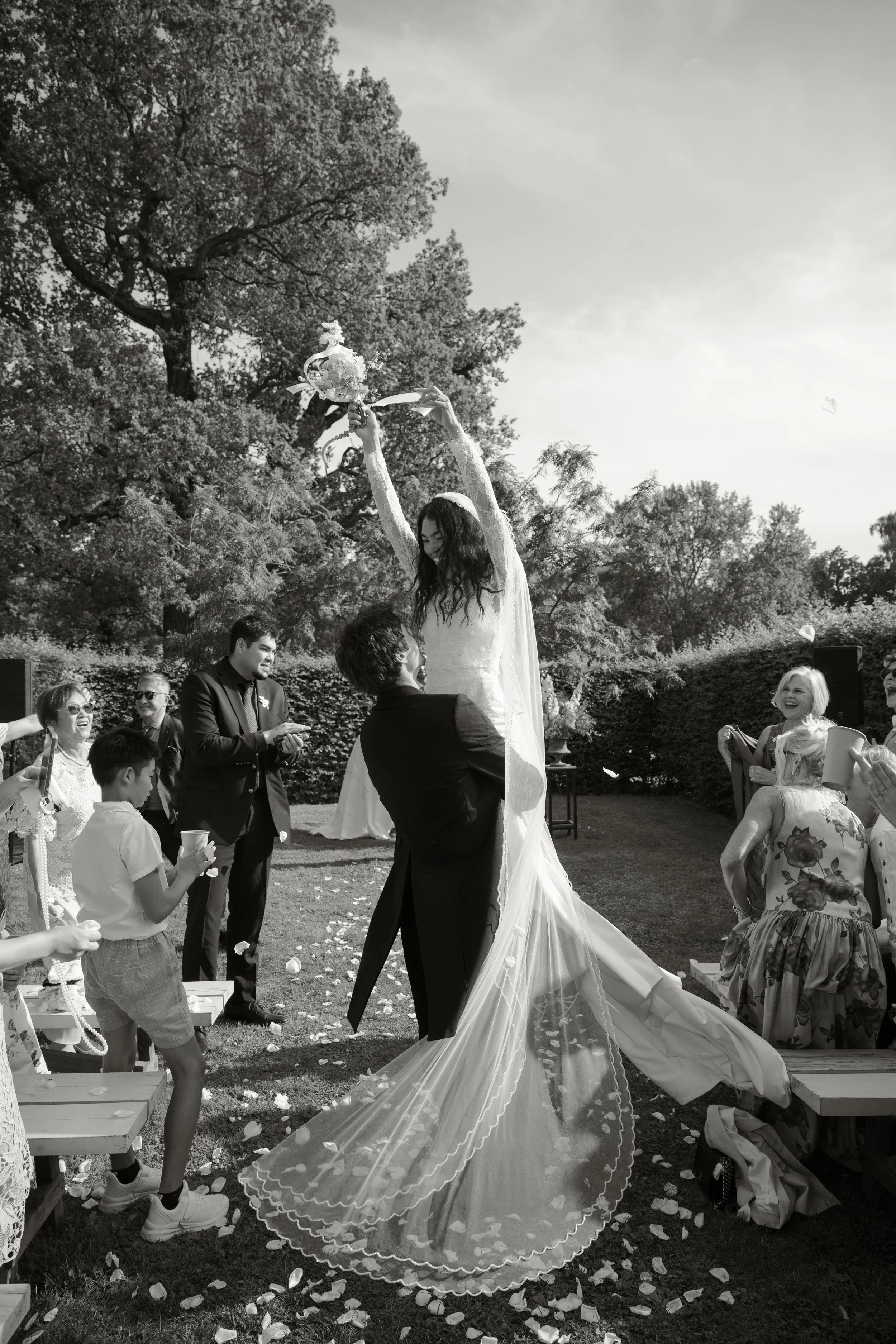 Bride being lifted by a man during wedding celebration outdoors, with guests celebrating and trees in background.