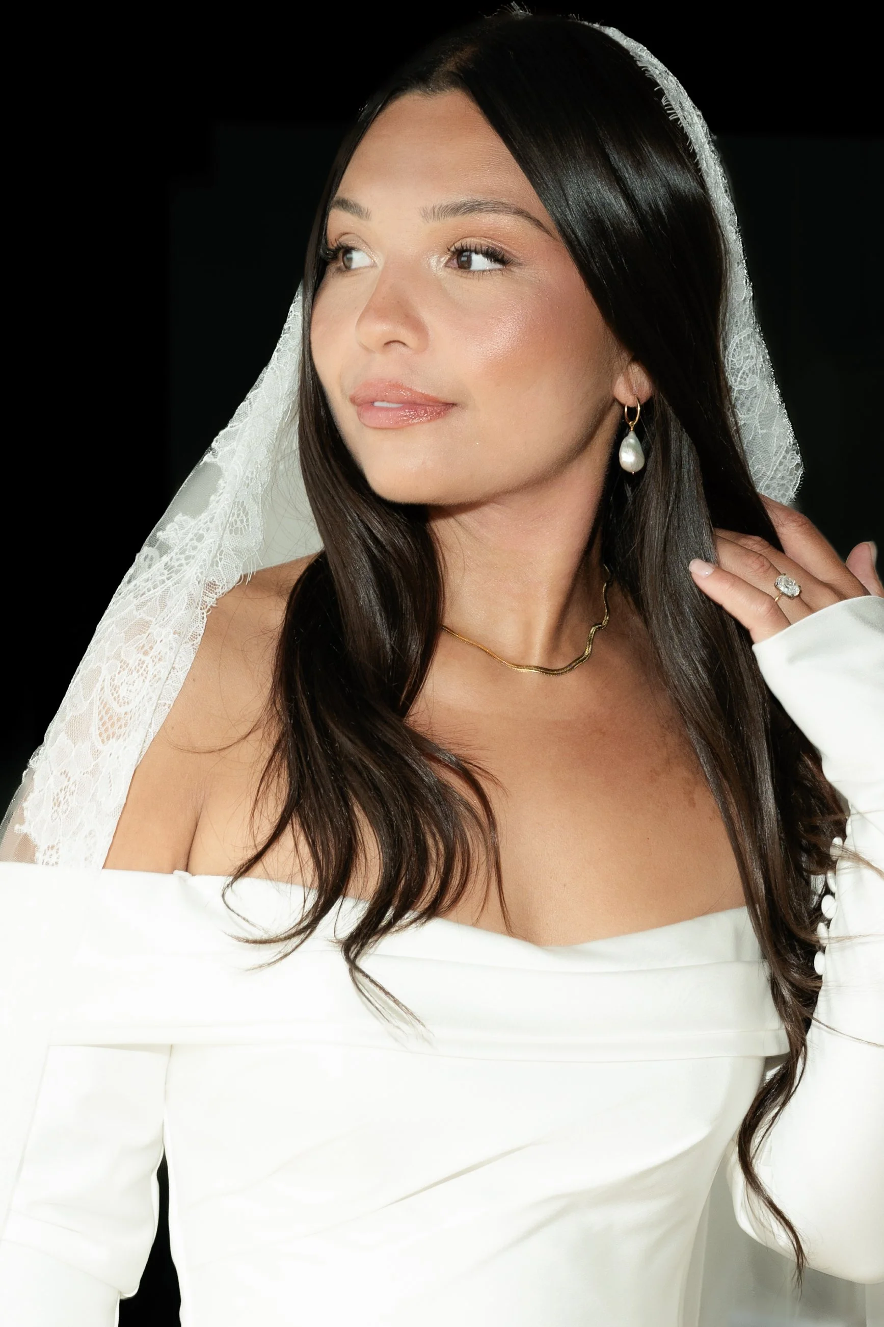 A woman in a white off-shoulder dress with a lace veil, wearing pearl earrings, a gold necklace, and a ring, posing against a black background.