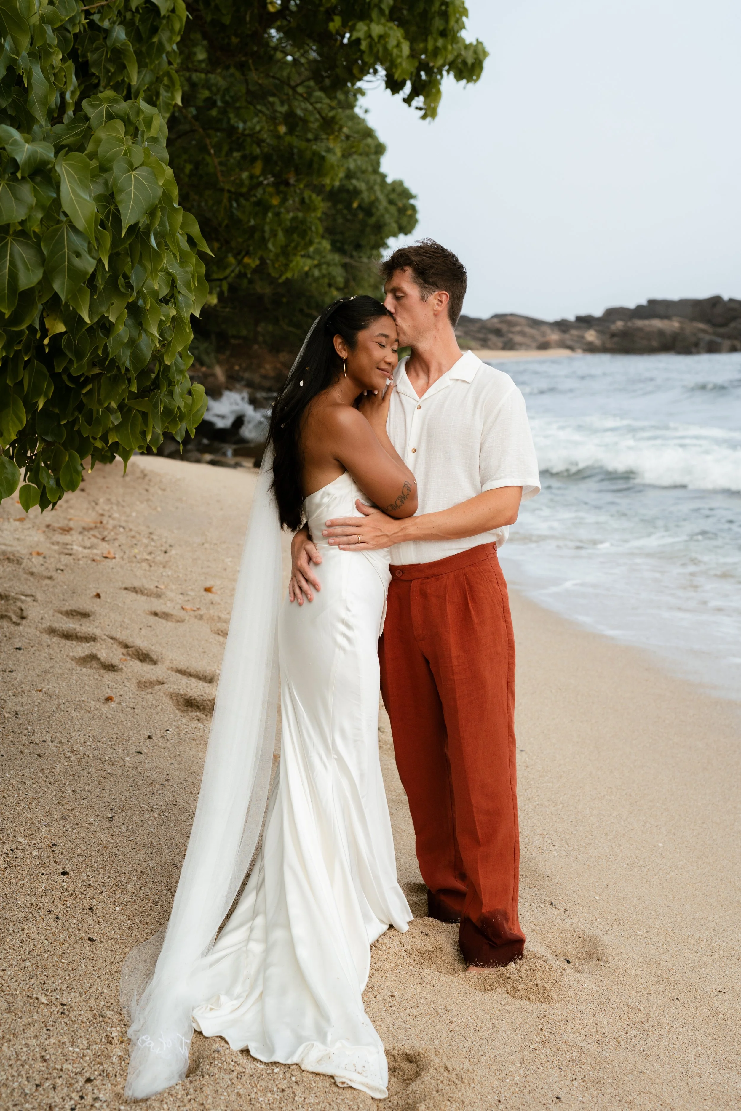 A couple dressed in wedding attire sharing a tender moment on the beach, with the ocean waves and greenery in the background.