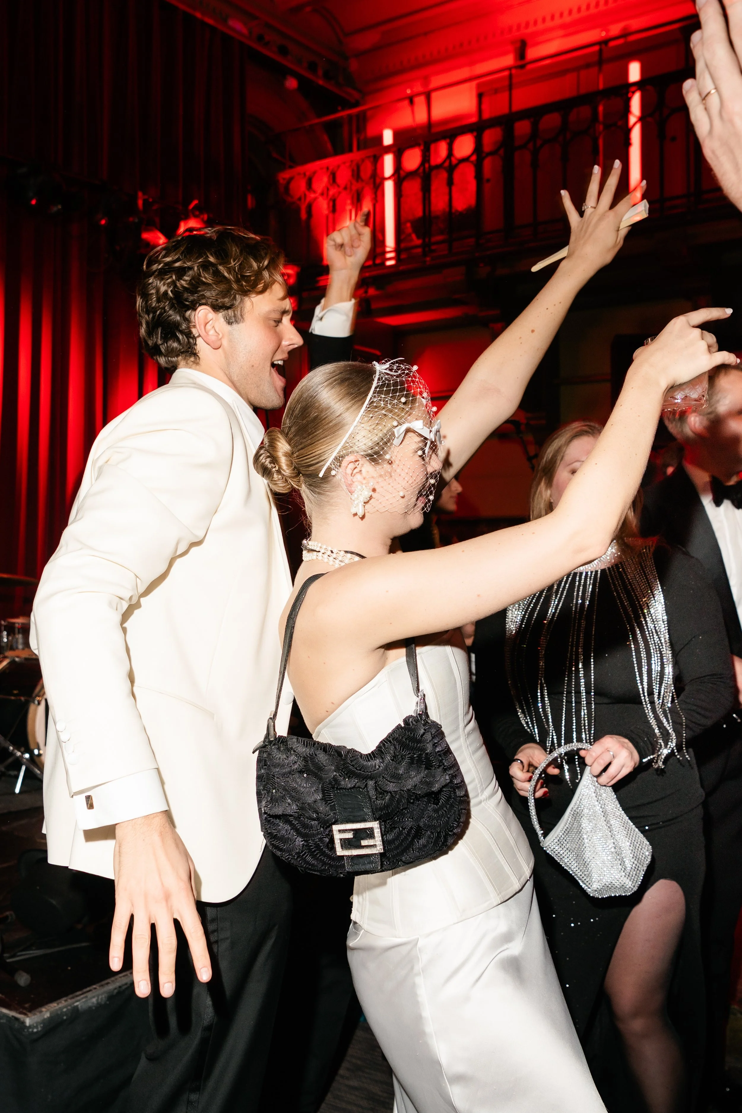 People dancing at a celebration or party in an elegant setting with red lighting; a woman in a white dress with a pearl necklace and a black handbag, and a man in a white tuxedo jacket and black pants, dancing and raising their arms.