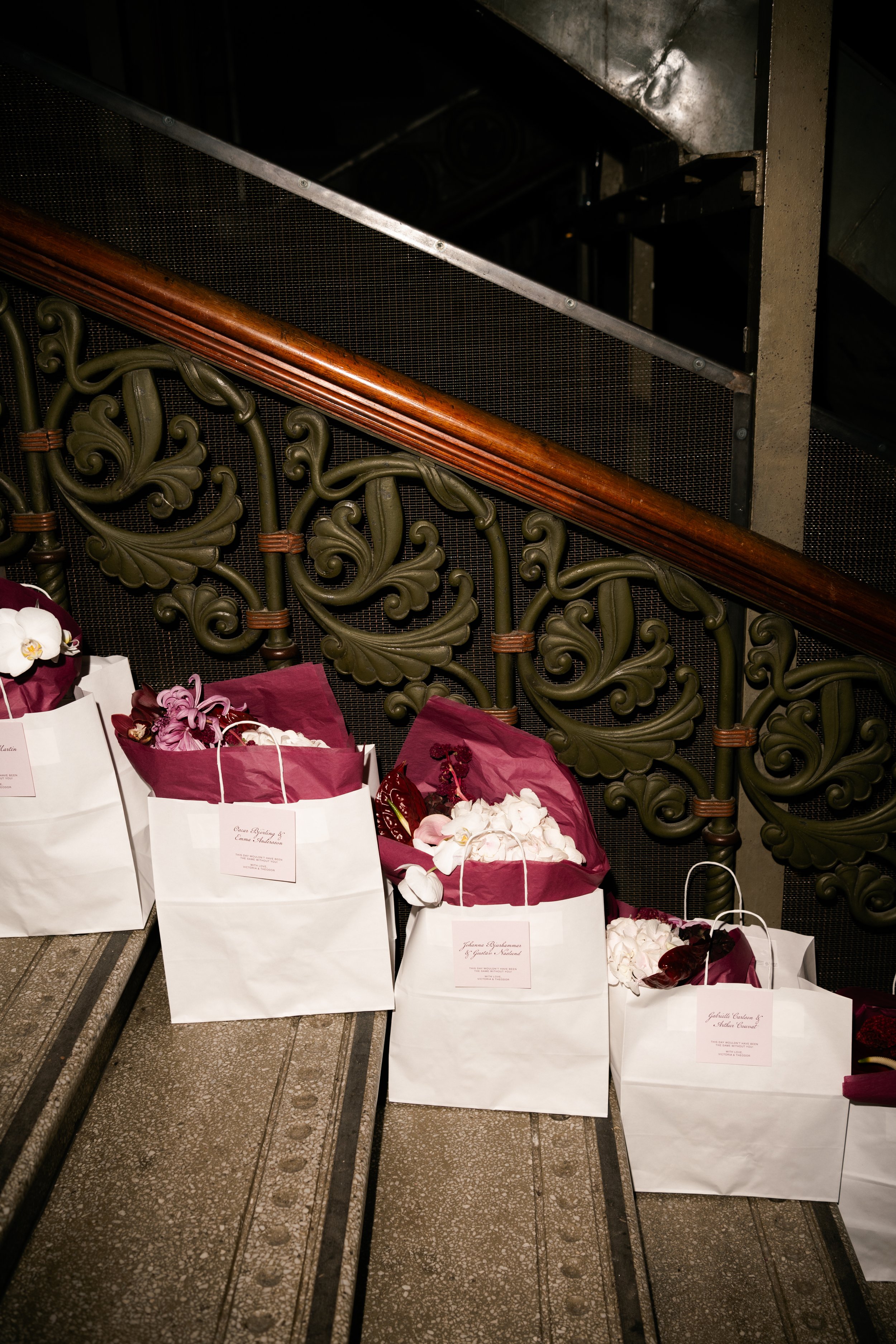 White shopping bags with pink tissue paper and flowers placed on the steps of a staircase with ornate green railings and a wooden handrail.