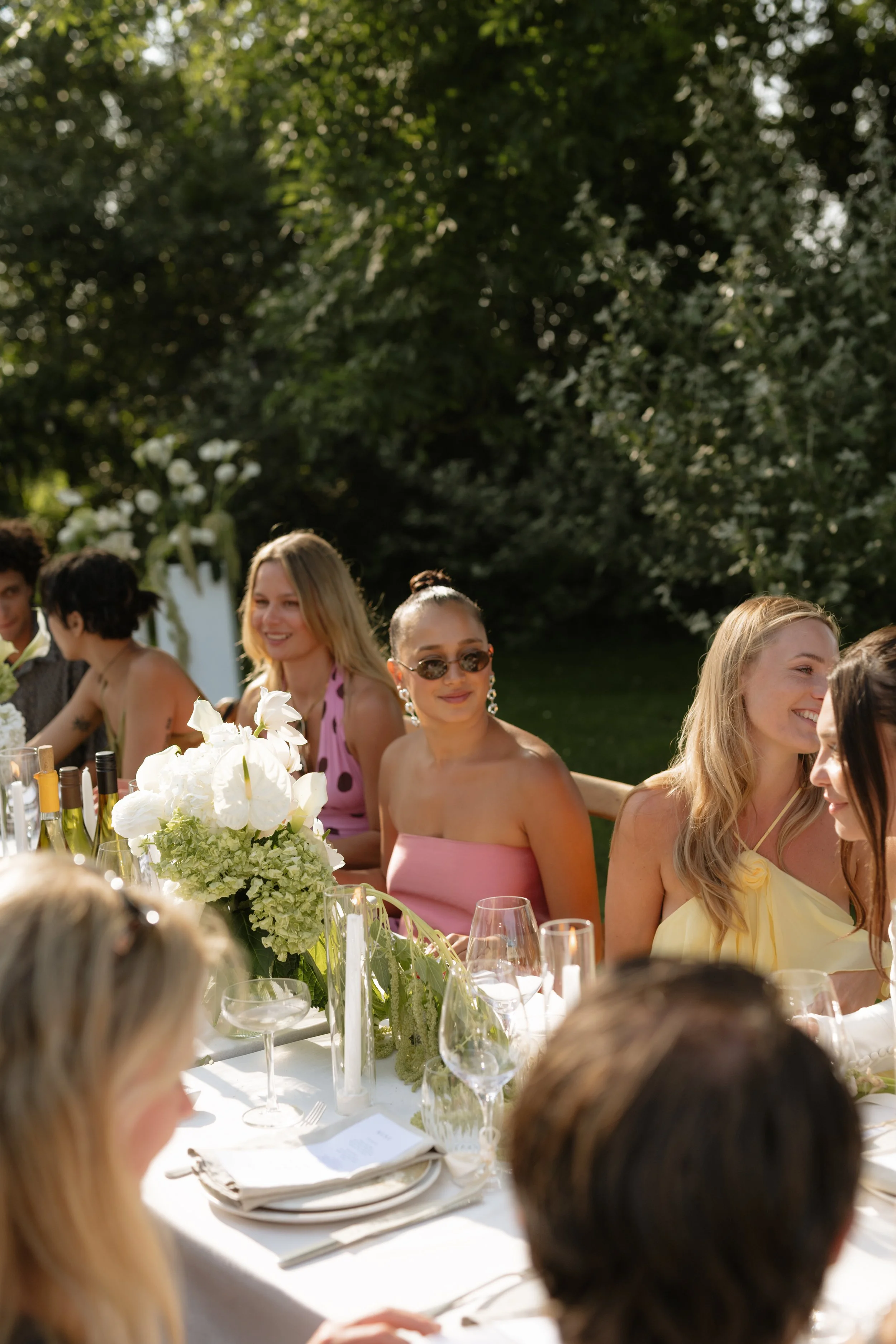 People sitting around a decorated outdoor dining table with flowers and wine bottles, enjoying a sunny day.