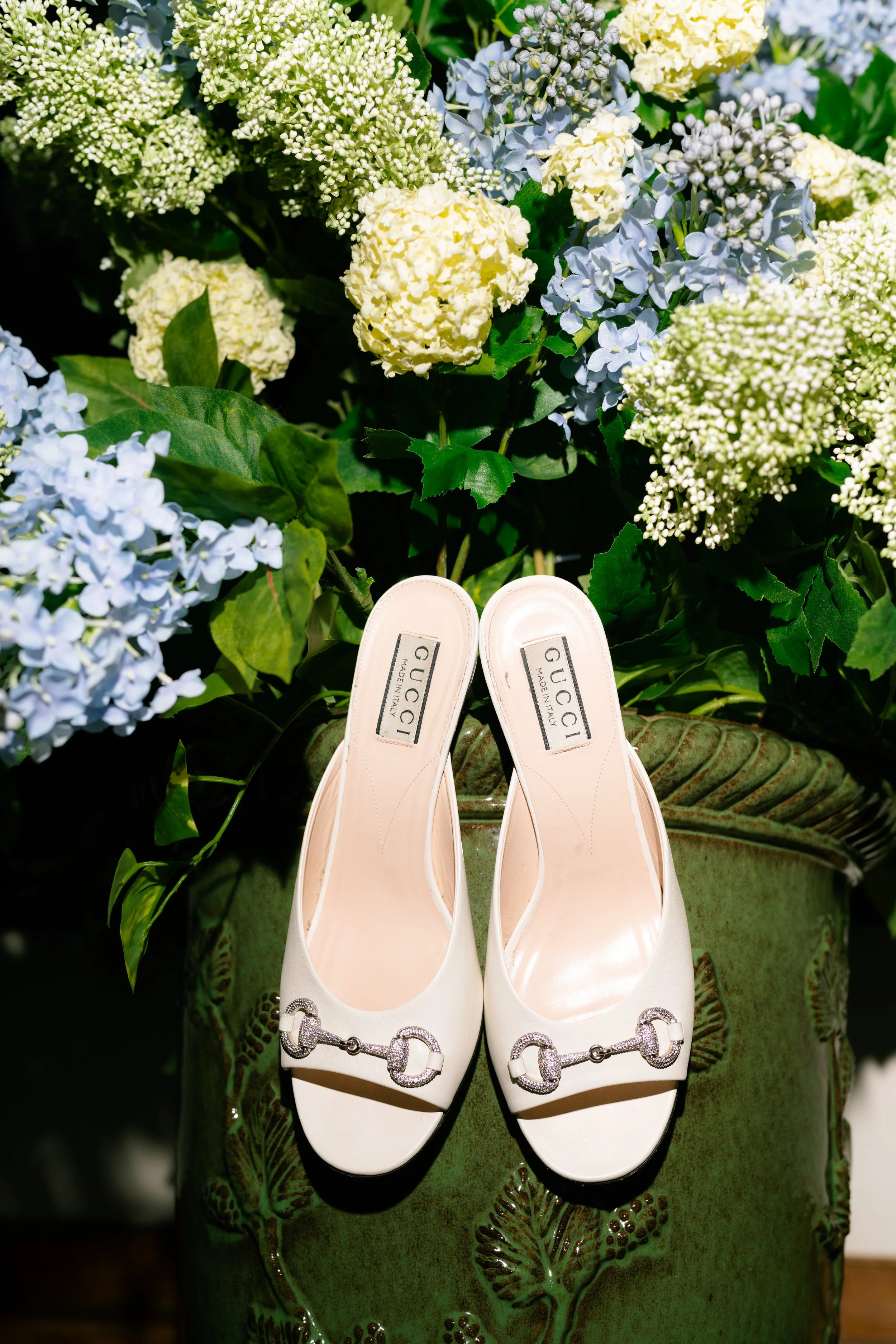 A pair of white Gucci high heel shoes with decorative buckles are placed on a green ceramic planter, with a background of white, yellow, and blue hydrangea flowers and green leaves.